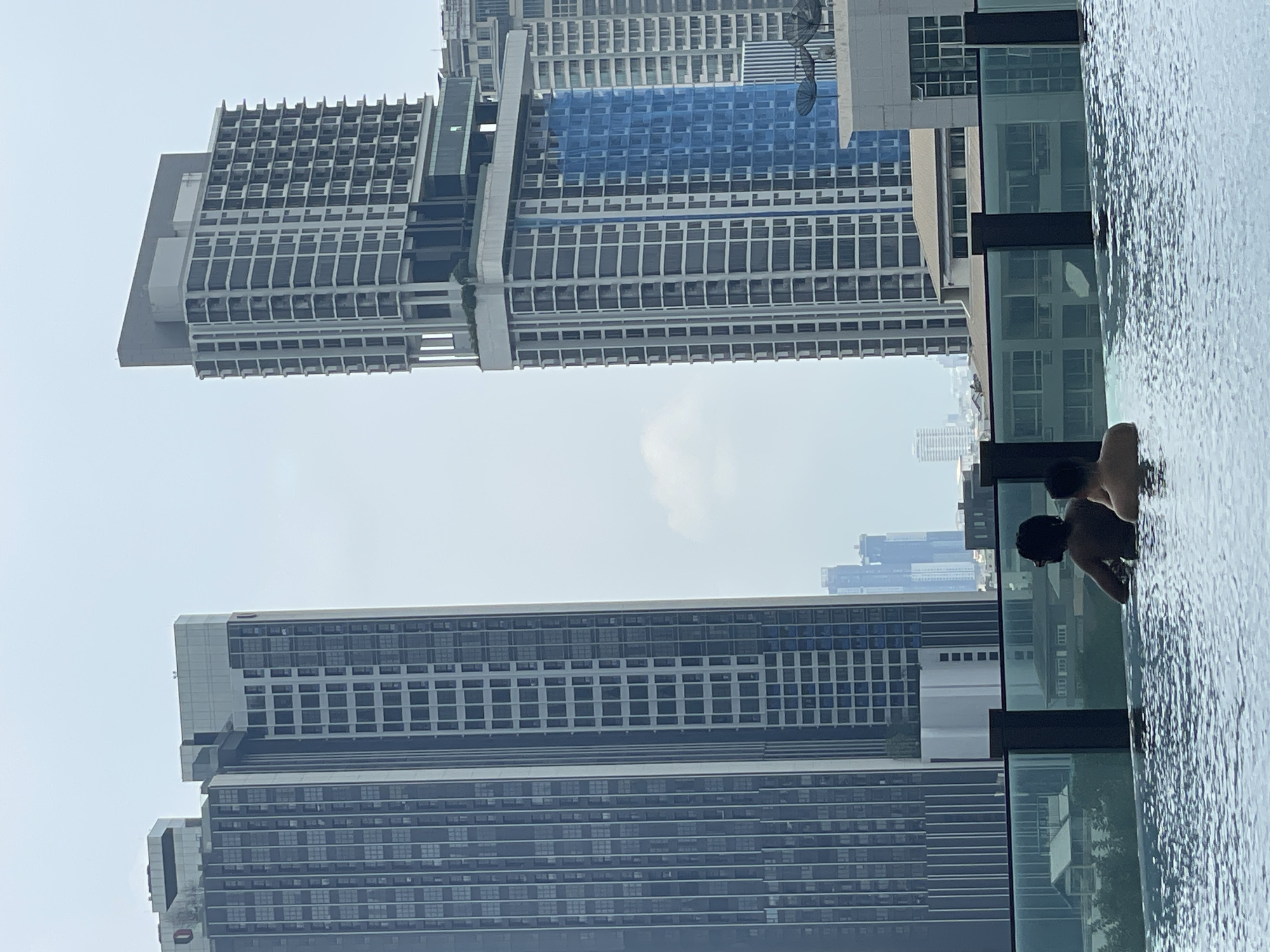 This picture shows a partly cloudy Bangkok, Thailand, skyline from the perspective of a rooftop pool, where at least two people are swimming. Buildings are tall and modern. Some buildings are under construction with blue netting and construction equipment. The people in the pool are in the lower right quadrant of the picture. The water in the pool is slightly rippled.