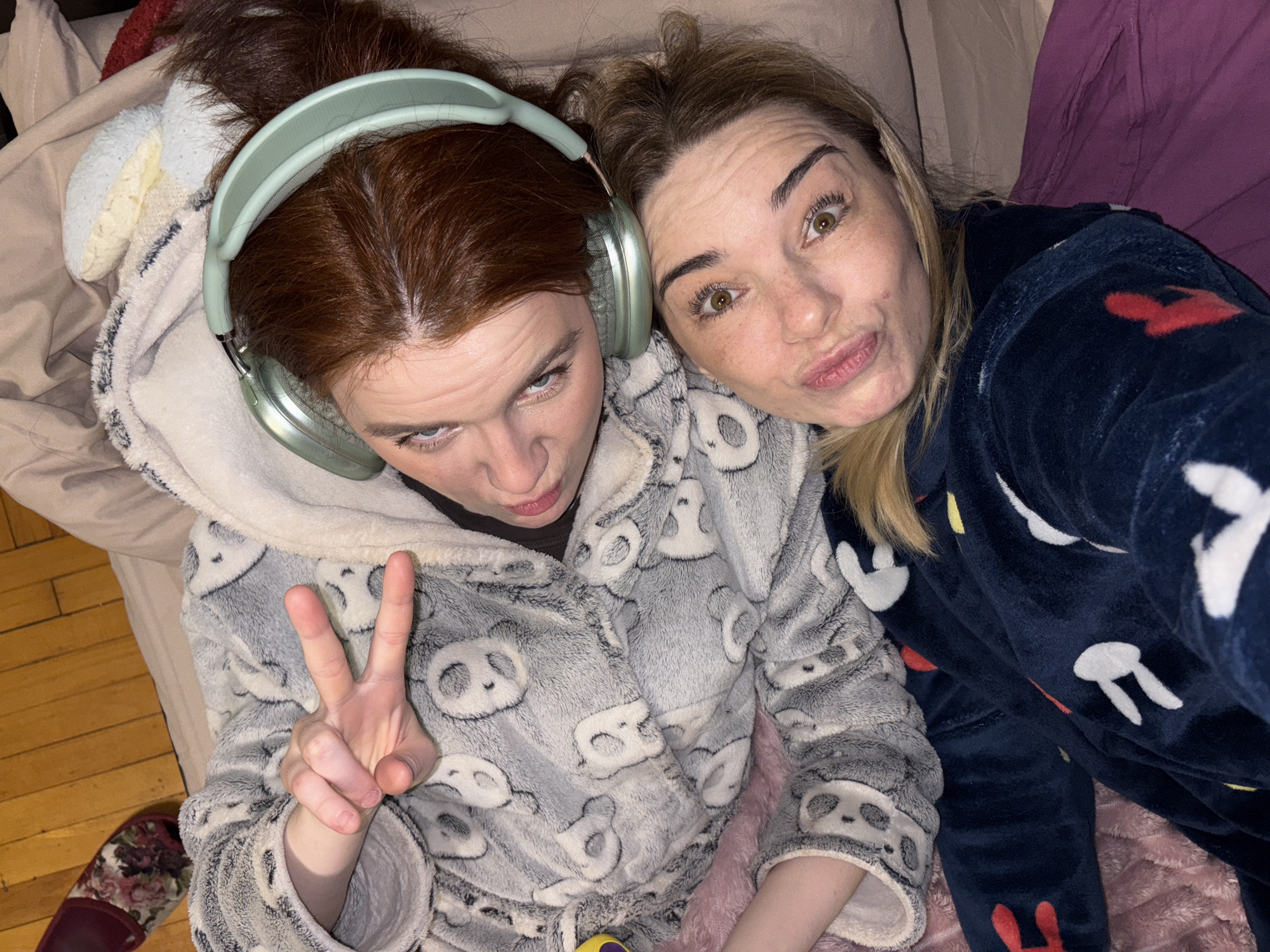 Two women lie on a bed in an indoor setting. One woman, with auburn hair pulled back, wears pale green headphones and a grey panda-patterned hooded robe, making a peace sign with her fingers. The second woman, with blonde highlights, is dressed in a dark blue bunny-patterned robe, making a kiss face towards the camera. Pillows and bedding are visible beneath them. The environment includes glimpses of a hardwood floor. The location is Unknown, Unknown.