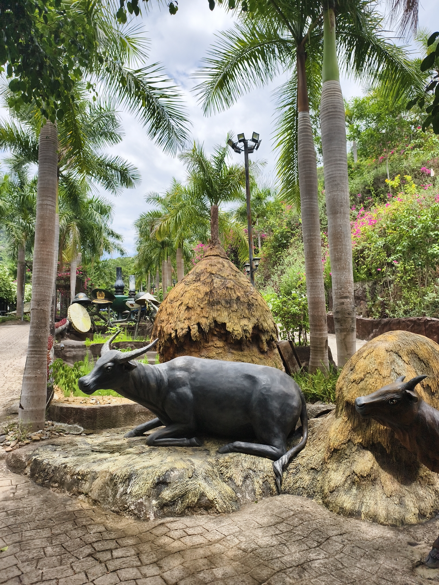 The image shows a park scene in Tay Nha Trang Ward, Vietnam, featuring life-size statues of water buffalo next to structures resembling haystacks. The statues are made of a dark material, possibly metal or painted concrete, and are positioned on a rock-like base. Tall palm trees line the park path, and lush greenery, including flowering bushes, fills the background. A lamp post is visible in the mid-ground, and a collection of vintage objects, including hats and oil lamps, can be seen further down the path. The ground is paved with interlocking bricks. The sky is overcast.