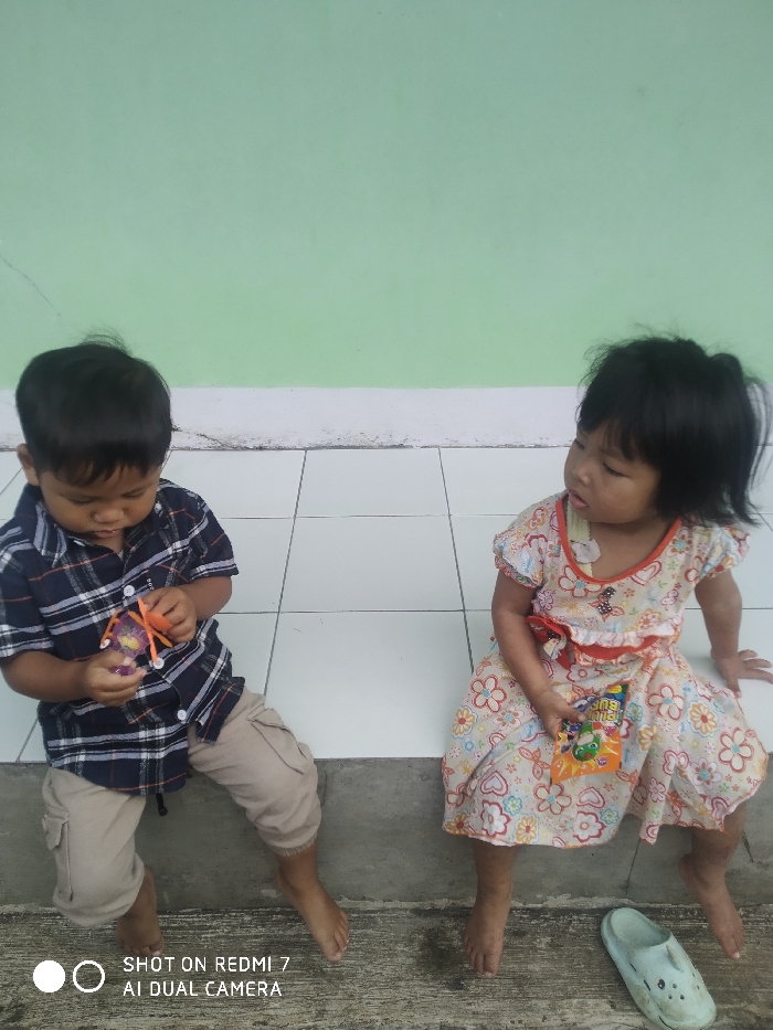 A young boy wearing a plaid shirt and khaki pants sits next to a young girl in a floral dress. Both children are barefoot and holding candy. They are seated on a ledge in front of a light green wall with white tile. A single white shoe lies on the ground next to the girl. The children appear to be focused on their candy.