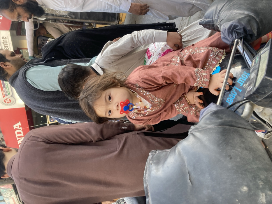A toddler is positioned on the front seat of a black motorcycle in an urban street environment within Quetta City Tehsil, Pakistan. The child, appearing to be approximately 1-2 years old, has light brown hair and wears a dusty rose-colored tunic adorned with silver-toned embroidery and sequins. A multi-strand beaded necklace is visible around the neck, and a red and blue pacifier is in the mouth. The child's right hand rests on the motorcycle's handlebars. A blue decal with the text "Your Lover" is visible on the motorcycle's fuel tank area.
Multiple individuals are present in the immediate vicinity. To the child's left, a man in a brown long-sleeved garment stands with his back partially to the camera. To the right and behind the child, a man in a light blue shirt and dark grey sleeveless vest is visible, looking downwards. Further to the right, a man with a white beard and glasses, dressed in a white shalwar kameez, faces towards the left.
In the background, a red sign with "HONDA" in white lettering is prominently displayed. Below it, white text indicates "3S SALES SERVICE SPARE PARTS". A yellow auto-rickshaw is partially visible on the right side of the frame. The scene indicates an active commercial area during daylight hours.