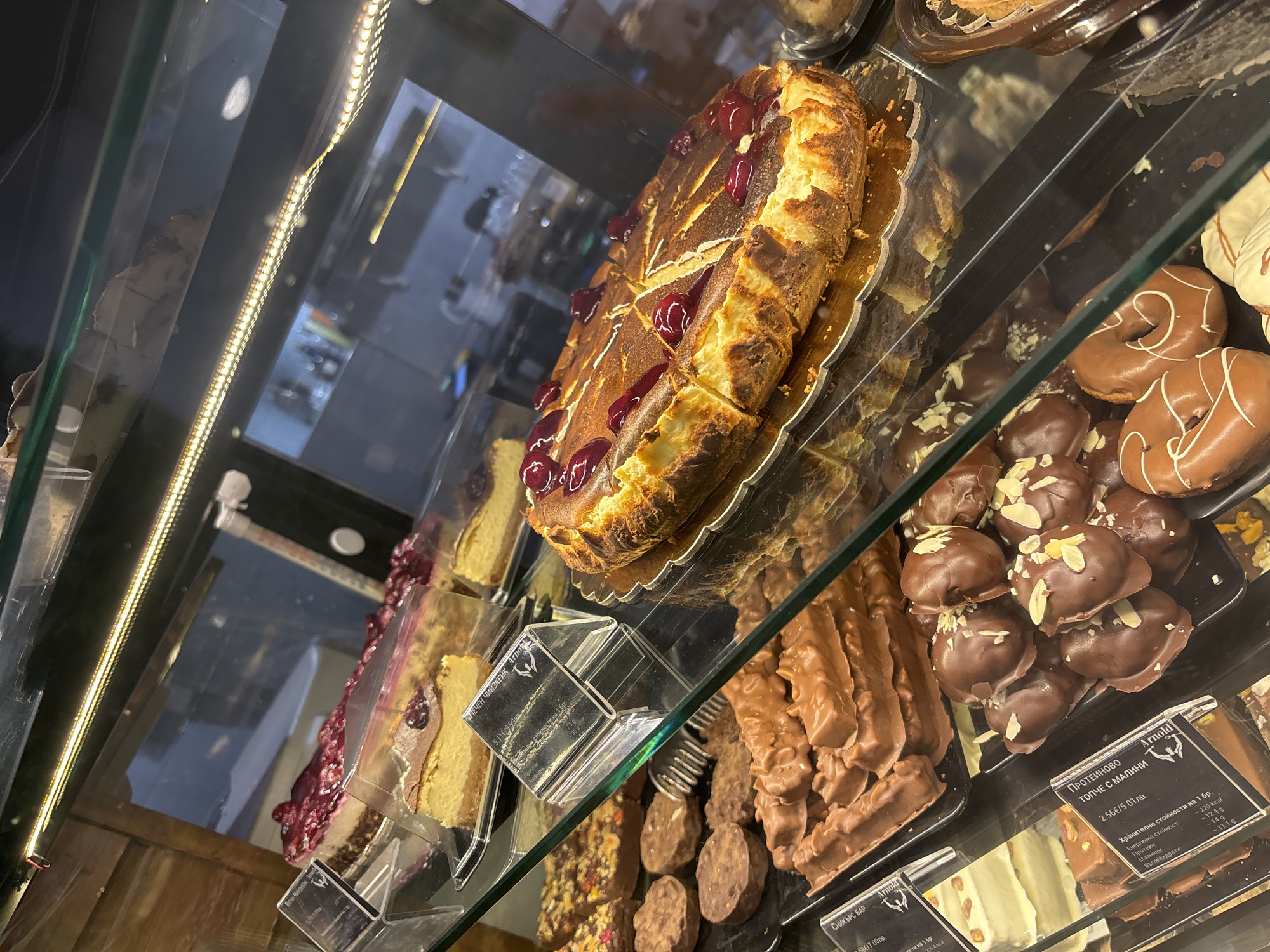 The image captures a display case filled with various desserts, potentially in a bakery or confectionery shop in Sofia, Bulgaria. A large, round cheesecake topped with red berries sits prominently on a gold-colored tray. Slices of a similar cake are visible nearby. To the right, there is a selection of chocolate-covered treats, including donuts with white chocolate drizzle and small, round chocolates with almond flakes. A sign indicates that some of the items are from "Arnold" and includes labels like "СНИКЪРС БАР", "ПРОТЕИНОВО ТОПЧЕ С МАЛИНИ", and "Веган Чийзкейк". These labels provide nutritional information in Bulgarian ("Хранителни стойности"). The environment suggests an indoor setting, likely a commercial establishment focused on baked goods and confectionery. The presence of LED lighting along the edge of the display case indicates a modern setting.