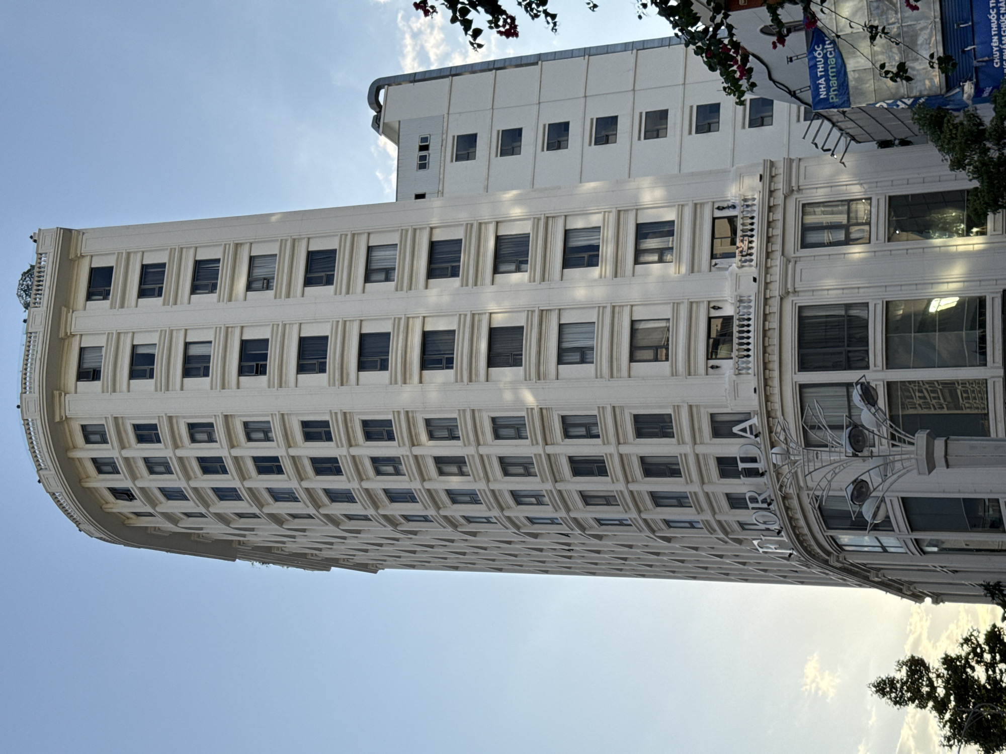 The image shows the exterior of a tall, white, multi-story building in Nha Trang, Vietnam, on a clear day with a blue sky and scattered clouds. The building has a curved facade with numerous windows and decorative architectural elements. The word "FLORIDA" is prominently displayed in large letters near the top of the building. A smaller, adjacent building is also visible to the right, featuring a sign that appears to be in Vietnamese and English, reading "NHÀ THUỐC Pharmacity". Trees and some flowering branches are present in the foreground and to the right of the buildings. The overall scene depicts an urban environment with a focus on the architecture of the buildings.