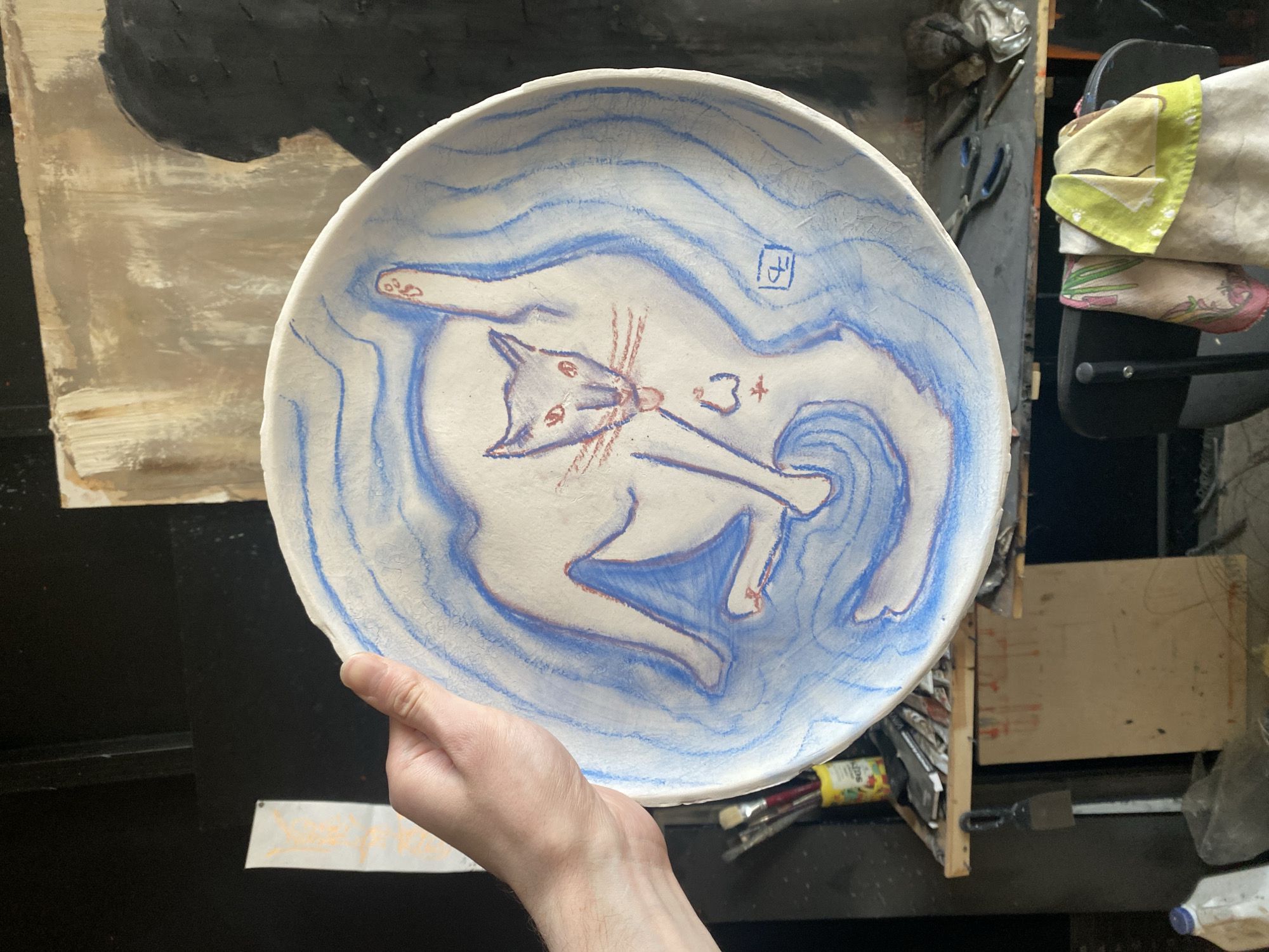 A person holds a handmade ceramic plate with a white cat design outlined in blue and red. The background is cluttered with art supplies and studio materials, suggesting an artist's workspace. There is no information in the image to indicate the location is in Moscow, Russia.