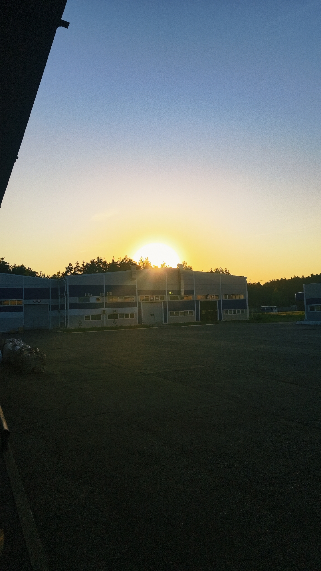 The image shows a low-angle, outdoor shot of a building at dusk. The building is a long, two-story structure with a blue and white exterior. There are numerous windows and several doors. The roofline is partially obscured by a line of trees. The sun is setting directly behind the building, casting a golden glow and creating lens flare. In the foreground, there's a dark, paved area that extends to the bottom of the frame. To the left, there's an area with bags of unidentified material. The location is Noviy, Russia.