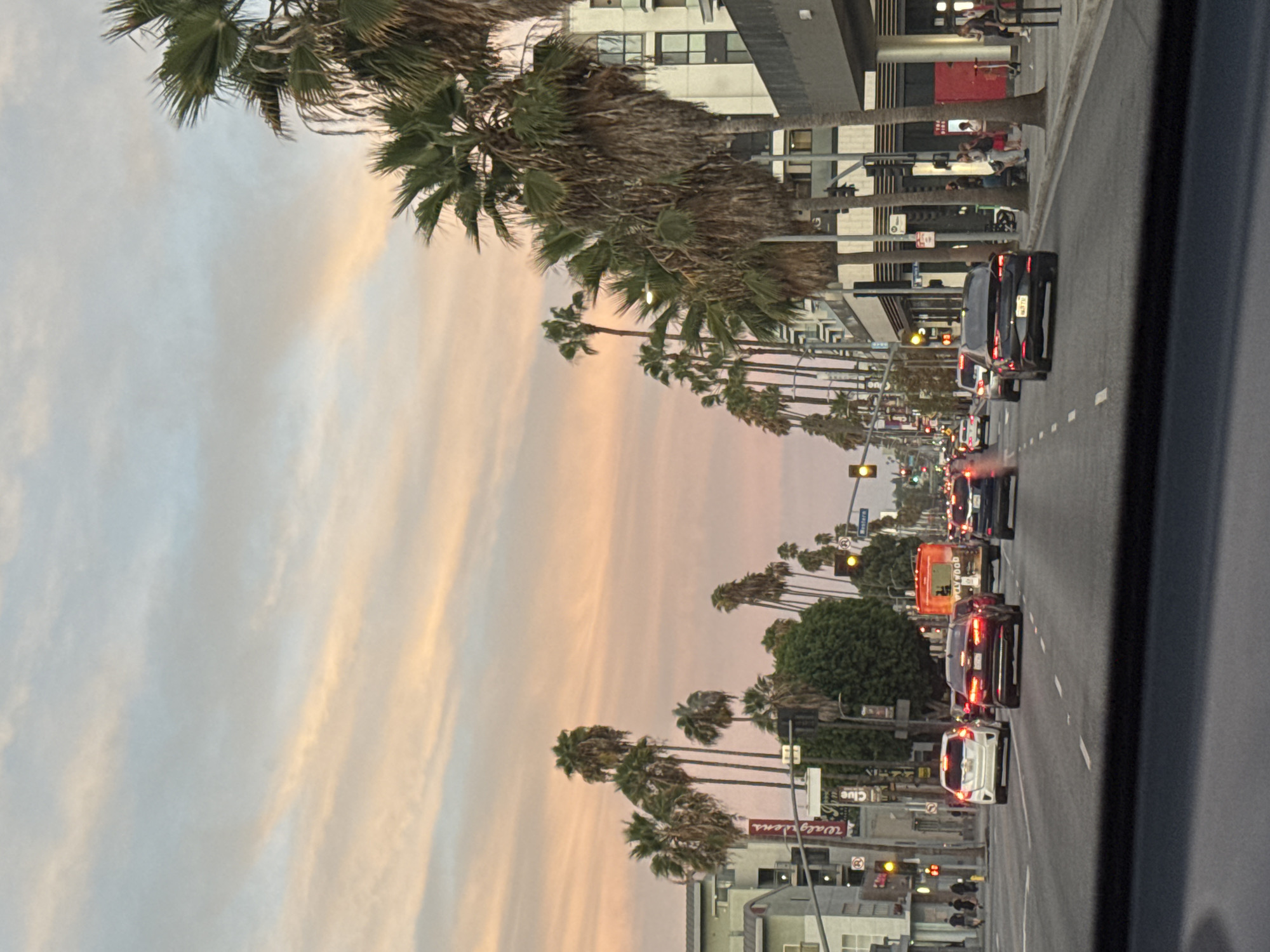The image depicts a street scene in Los Angeles, California, USA, during a sunset. The sky is filled with soft hues of pink, orange, and blue, indicative of twilight. The street is lined with palm trees on both sides, with buildings of varying architectural styles, including modern apartment buildings and older commercial structures. Traffic is present, with several cars and a bus visible on the road. Pedestrians are seen on the sidewalks, and some appear to be waiting at a bus stop. Traffic signals are illuminated, and streetlights are beginning to glow. The overall impression is of a busy urban environment transitioning into evening, with the characteristic palm trees and warm sky of Southern California.