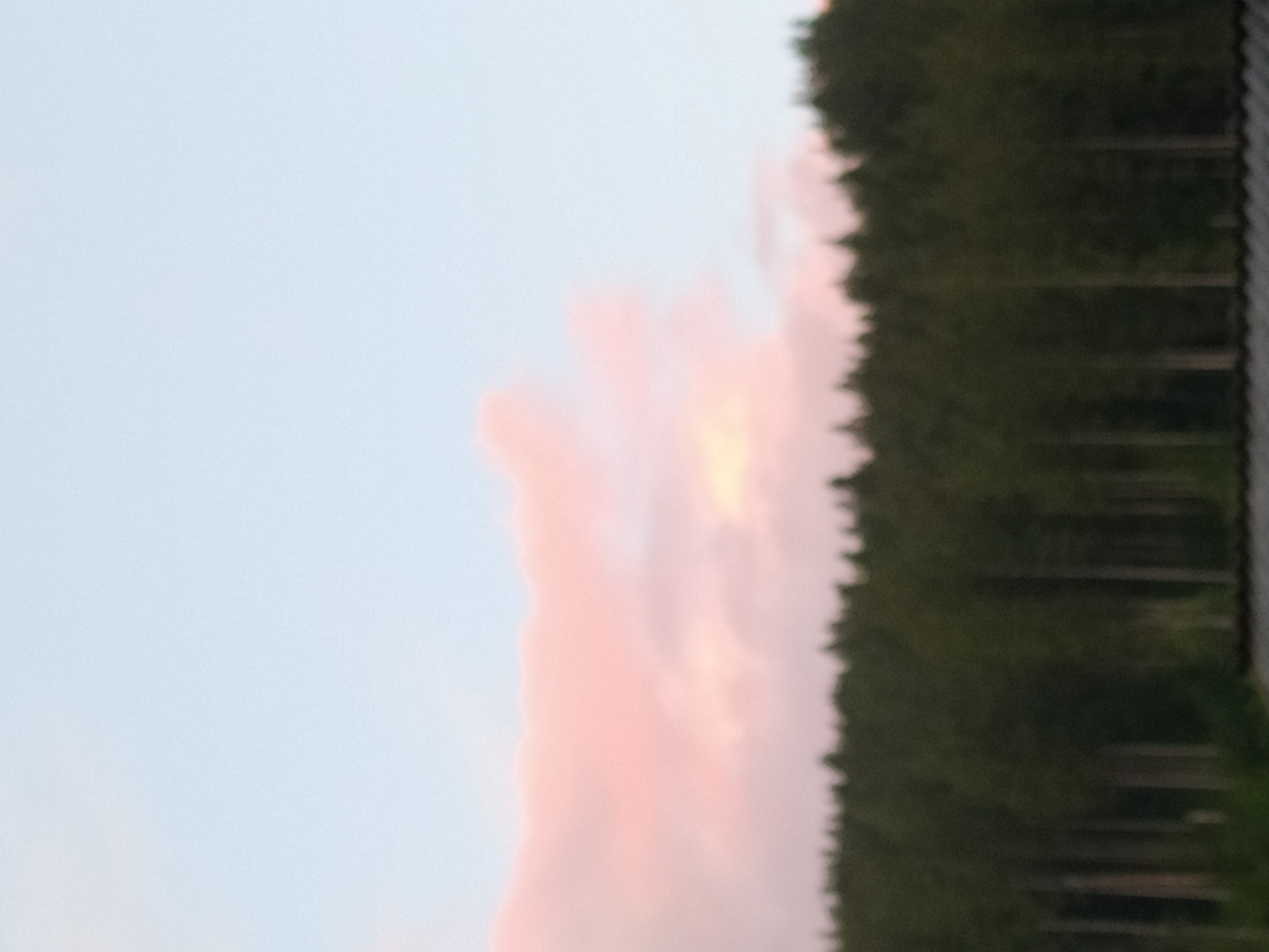 The image depicts a hazy, out-of-focus scene with a forest of dark green trees in the foreground and a pale blue sky with soft pink clouds at the top. The bottom edge of the image shows a blurry dark structure, possibly a roof. The scene appears to be an outdoor landscape, likely taken during twilight or dawn due to the coloration of the clouds. There are no visible people, animals, or specific identifiable structures beyond the indistinct dark object at the bottom. The image does not provide any specific visual cues to indicate its location within Hoverla, Ukraine, or any other city.