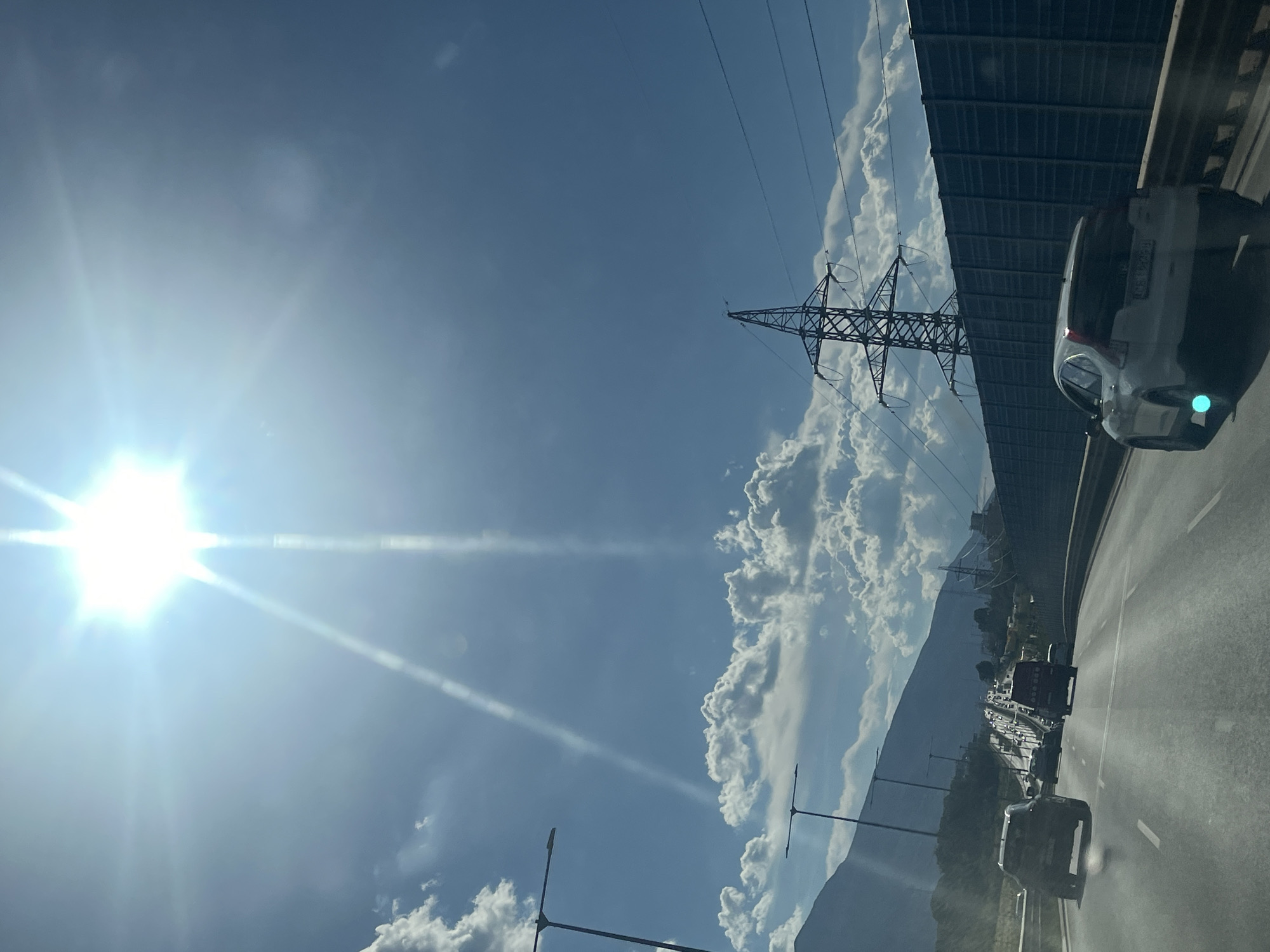 The image captures a bright, sunny day on a highway. The sun is prominently visible at the top of the frame, with intense light rays emanating from it, causing lens flares. The sky is a clear blue with scattered white clouds. A mountain range is visible in the background to the left, partially obscured by clouds.
On the highway, several cars are driving. In the foreground, a white SUV is in the right lane, and a dark-colored SUV is in the left lane. Further ahead, a line of cars and trucks is visible, indicating moderate traffic.
To the right of the highway, a tall sound barrier, likely made of dark panels, runs along the road. Behind the sound barrier, a large electricity pylon with several power lines is visible. Streetlights are also present along the side of the highway.
The overall scene suggests a typical highway journey on a sunny day, possibly in a mountainous region, with urban infrastructure like sound barriers and power lines present. The location is unknown.