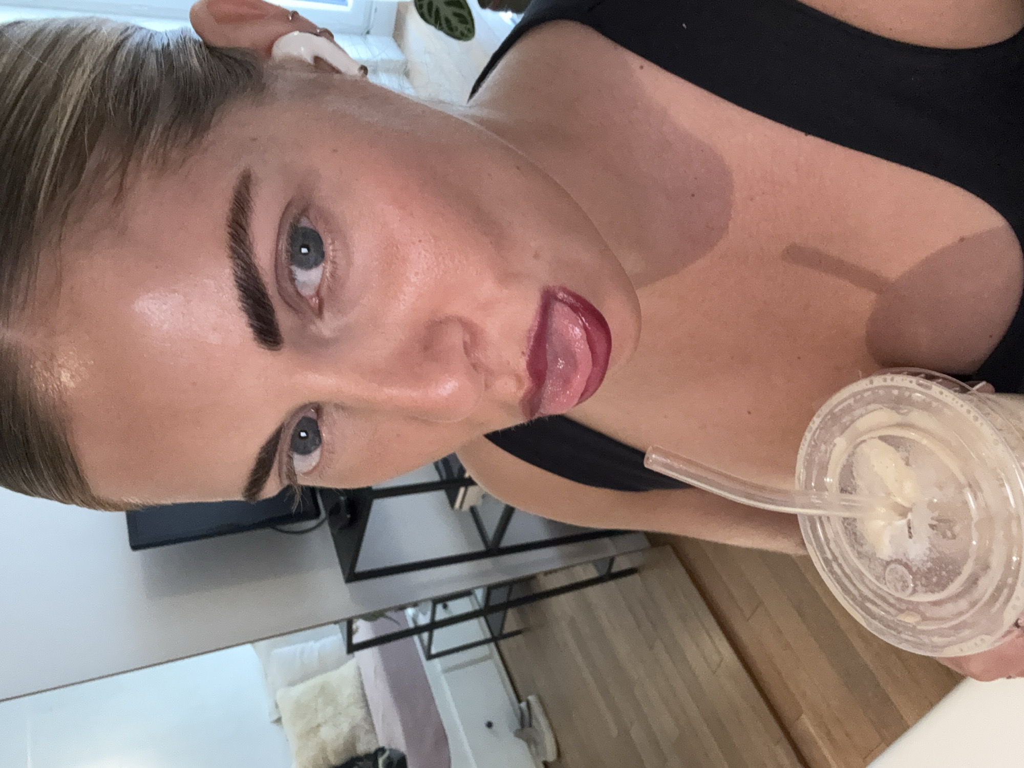A woman with blonde hair and fair skin is holding a clear plastic cup with a straw, possibly containing a beverage. She is wearing a black top. Her tongue is slightly out, and she is looking directly at the camera with blue eyes. The background includes a TV on a black stand, a multi-tiered shelving unit, and part of a light-colored floor and wall. The location does not appear to be in Paris, France.