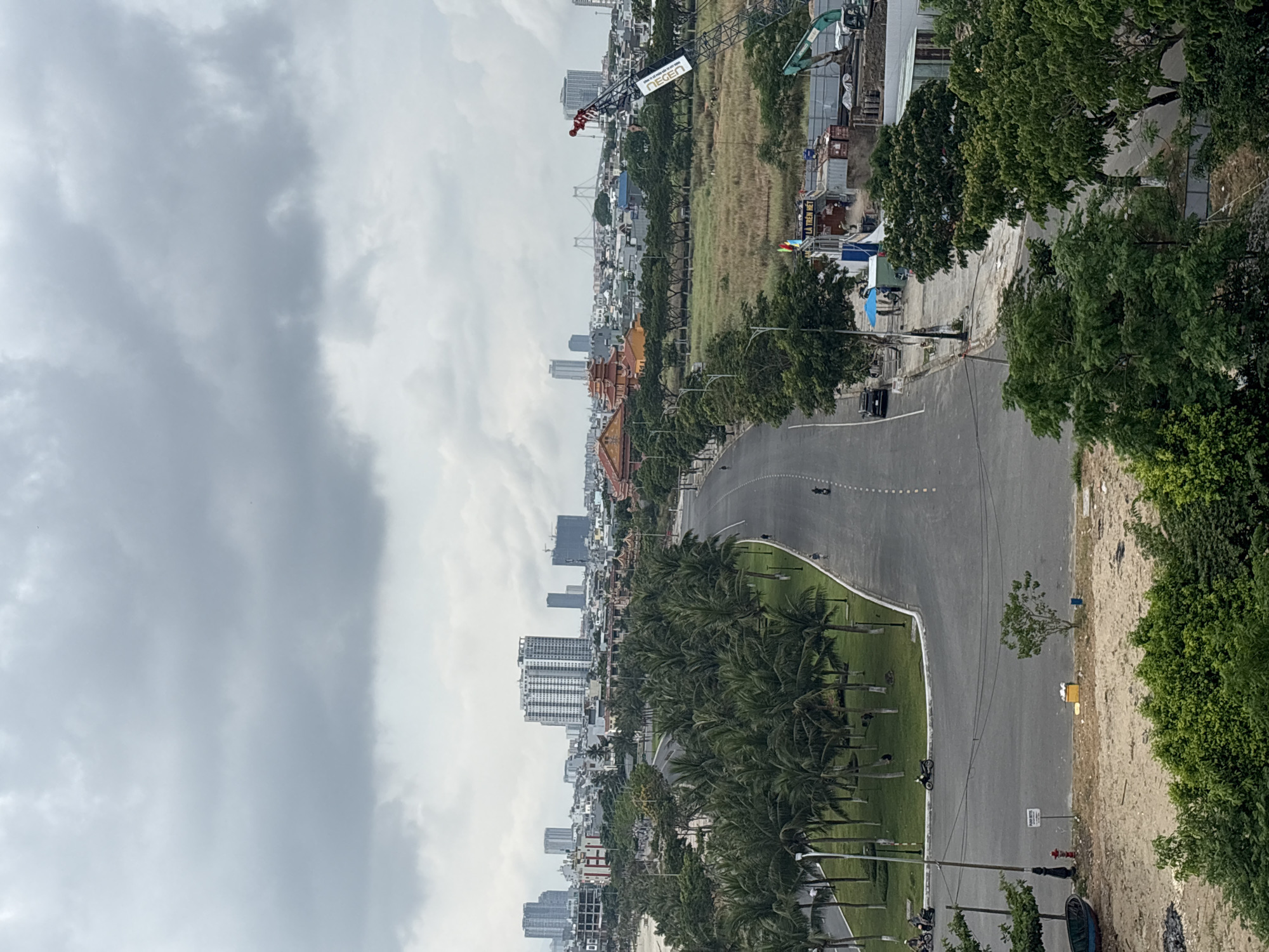 The image depicts a cityscape in Đà Nẵng, Vietnam, from an elevated perspective. The foreground features a multi-lane road running parallel to the coast, lined with palm trees. In the background, there are tall buildings, a bridge, and a large open field with construction activity. The sky is overcast, and the lighting is subdued. A few motorbikes are visible on the road.
