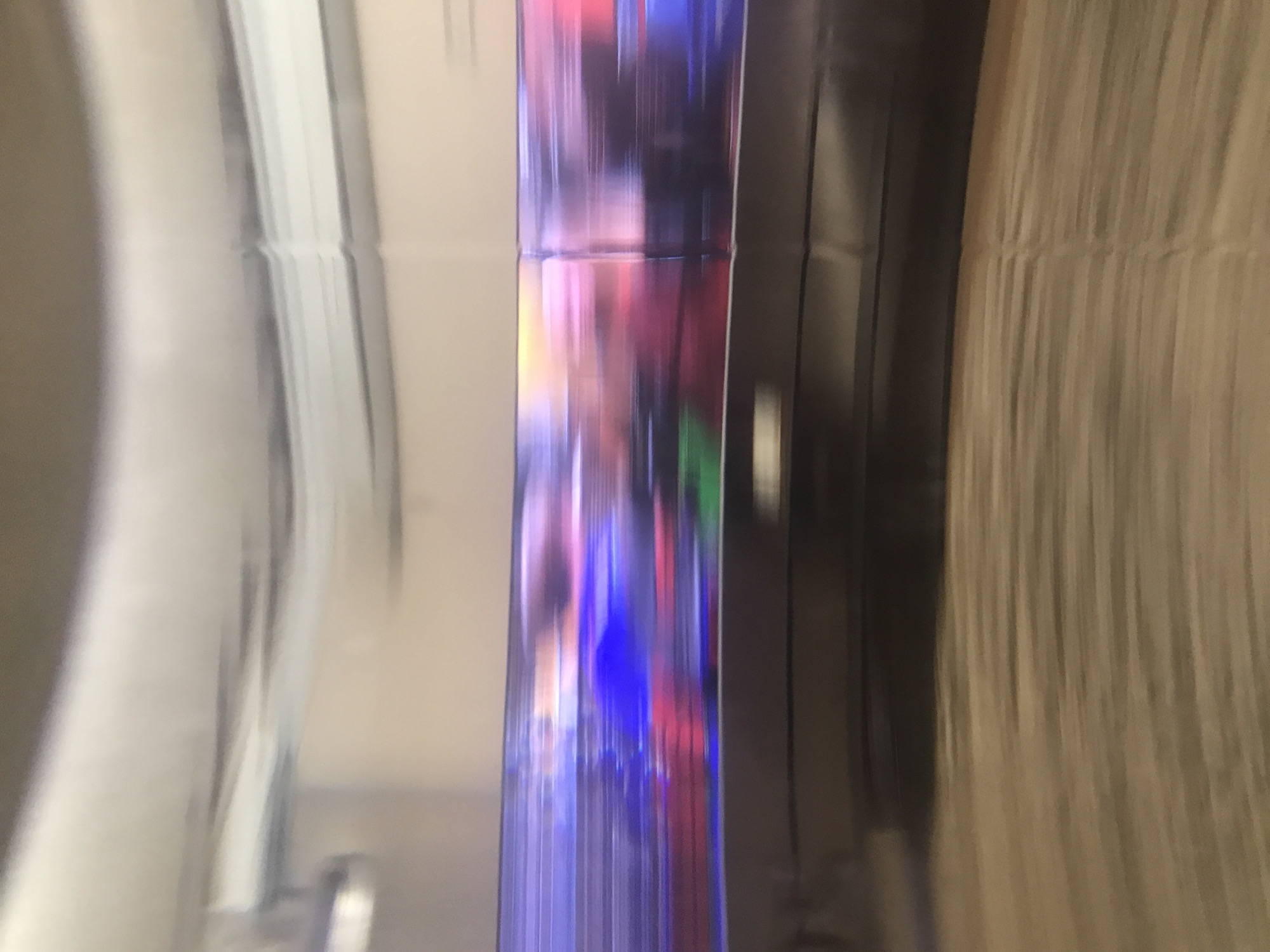 The image depicts a distorted reflection of an urban scene, possibly in Yerevan, Armenia. The reflection is visible on a vertical, shiny surface, likely glass or polished metal. The reflected scene contains a blur of colors, suggesting city lights, moving vehicles, or signage. The reflection is segmented, potentially indicating that it is reflected on multiple panels. To the left of the reflective surface is a blurred, light-colored area with vertical lines. To the right is a darker surface with vertical patterns, which might be curtains or another wall. There are no people directly visible in the reflection or the surrounding surfaces. The scene is indoors, as indicated by the presence of the wall and curtains.
