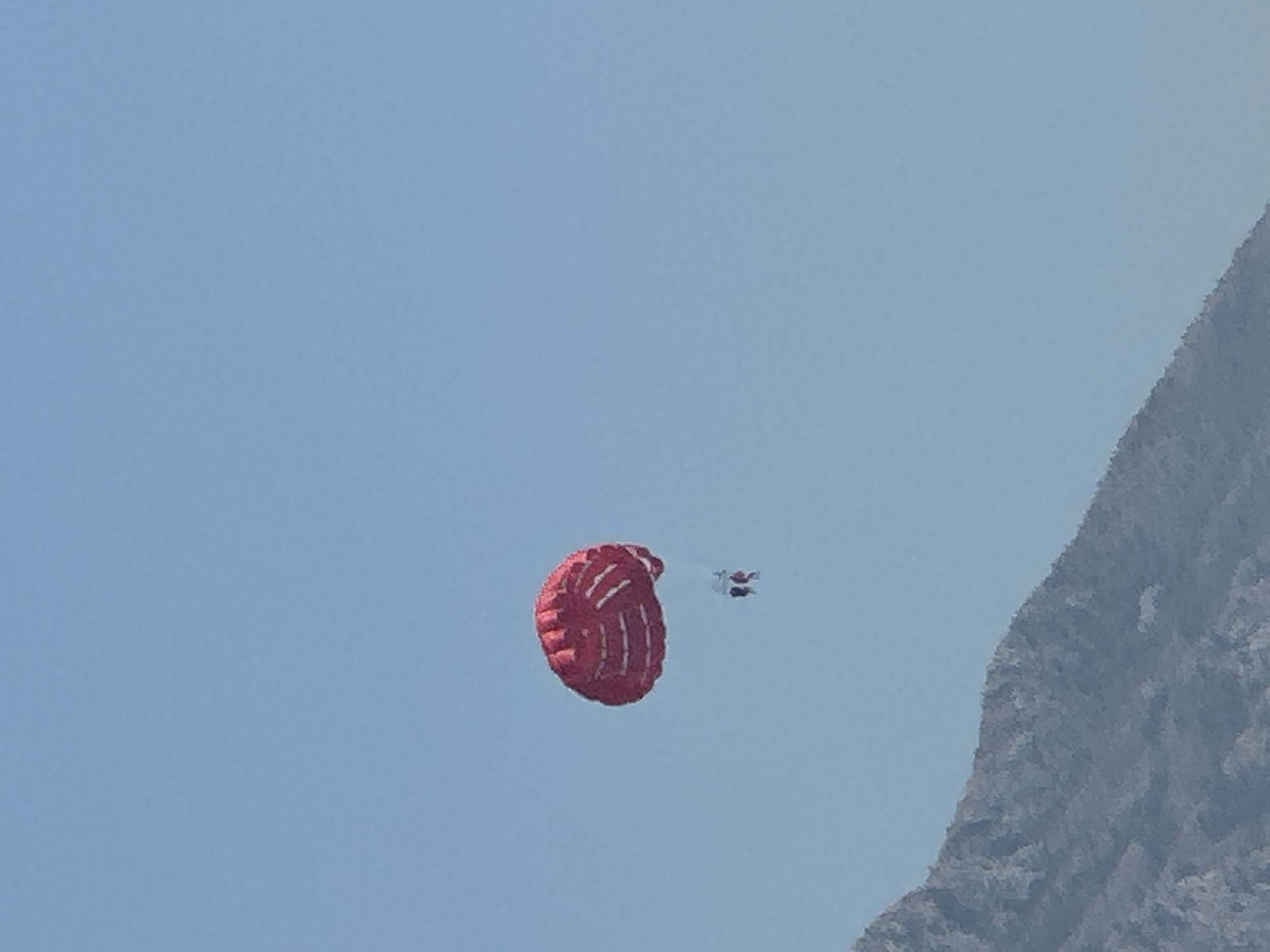 A red and white striped parachute is visible in a clear blue sky. Two individuals are suspended beneath the parachute, appearing to be parasailing. In the lower left portion of the image, a rugged, rocky mountain slope is present, partially obscuring the view. The scene is consistent with an outdoor recreational activity.