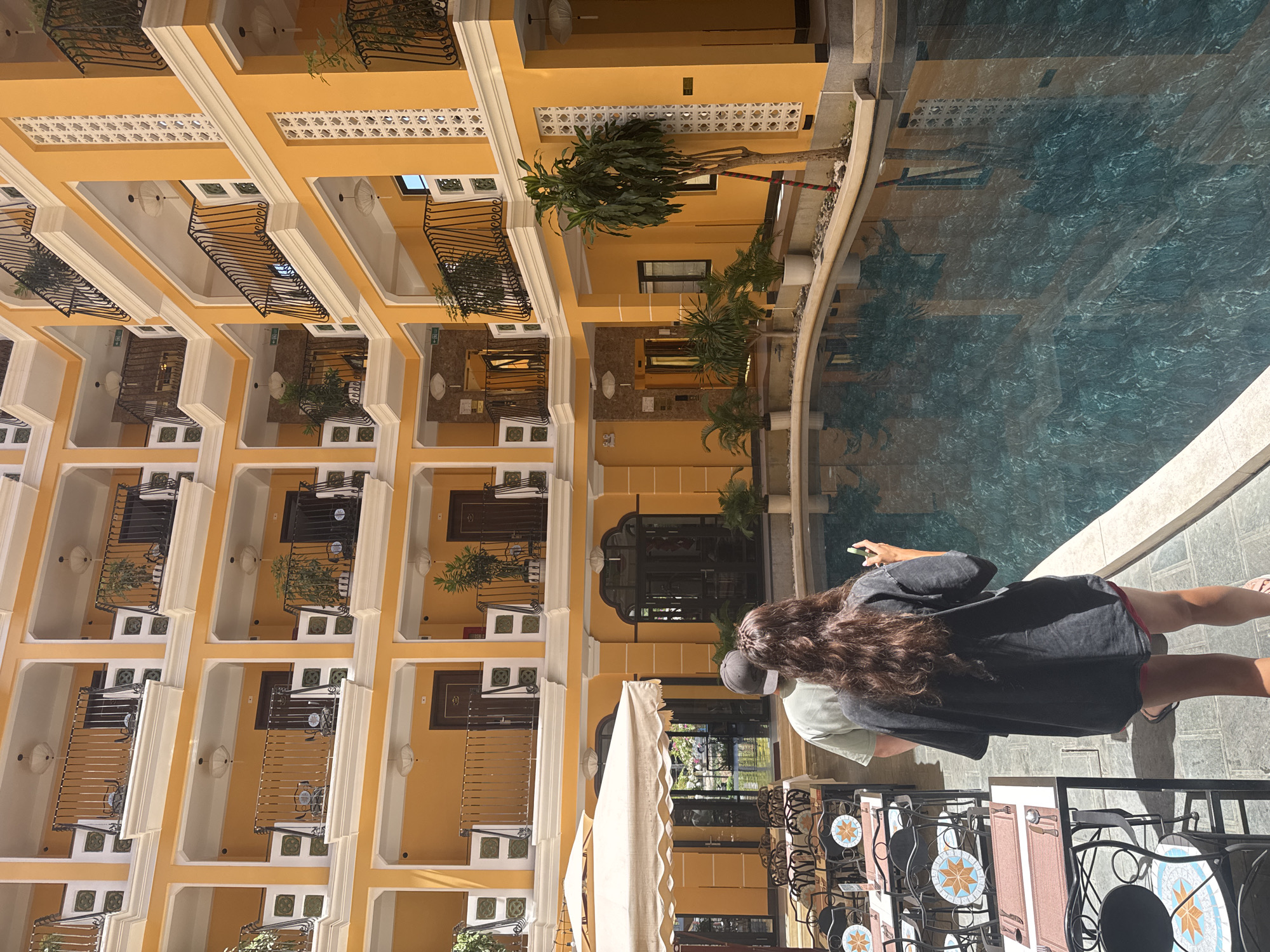 The image shows a courtyard scene in what appears to be a hotel or resort, possibly located in Đà Nẵng, Vietnam. The buildings are multi-story, with yellow walls, white trim, and balconies featuring black metal railings. Some of the balconies contain small potted plants. A turquoise swimming pool is visible along one side of the courtyard, reflecting the architecture. Two people are standing near the pool, a person with long curly brown hair wearing a dark t-shirt and a light-colored shirt. There are tables and chairs in the foreground, suggesting a dining or relaxation area, possibly part of a restaurant or café. A partially visible white awning covers some of the tables. The perspective is from slightly above, looking down into the courtyard.