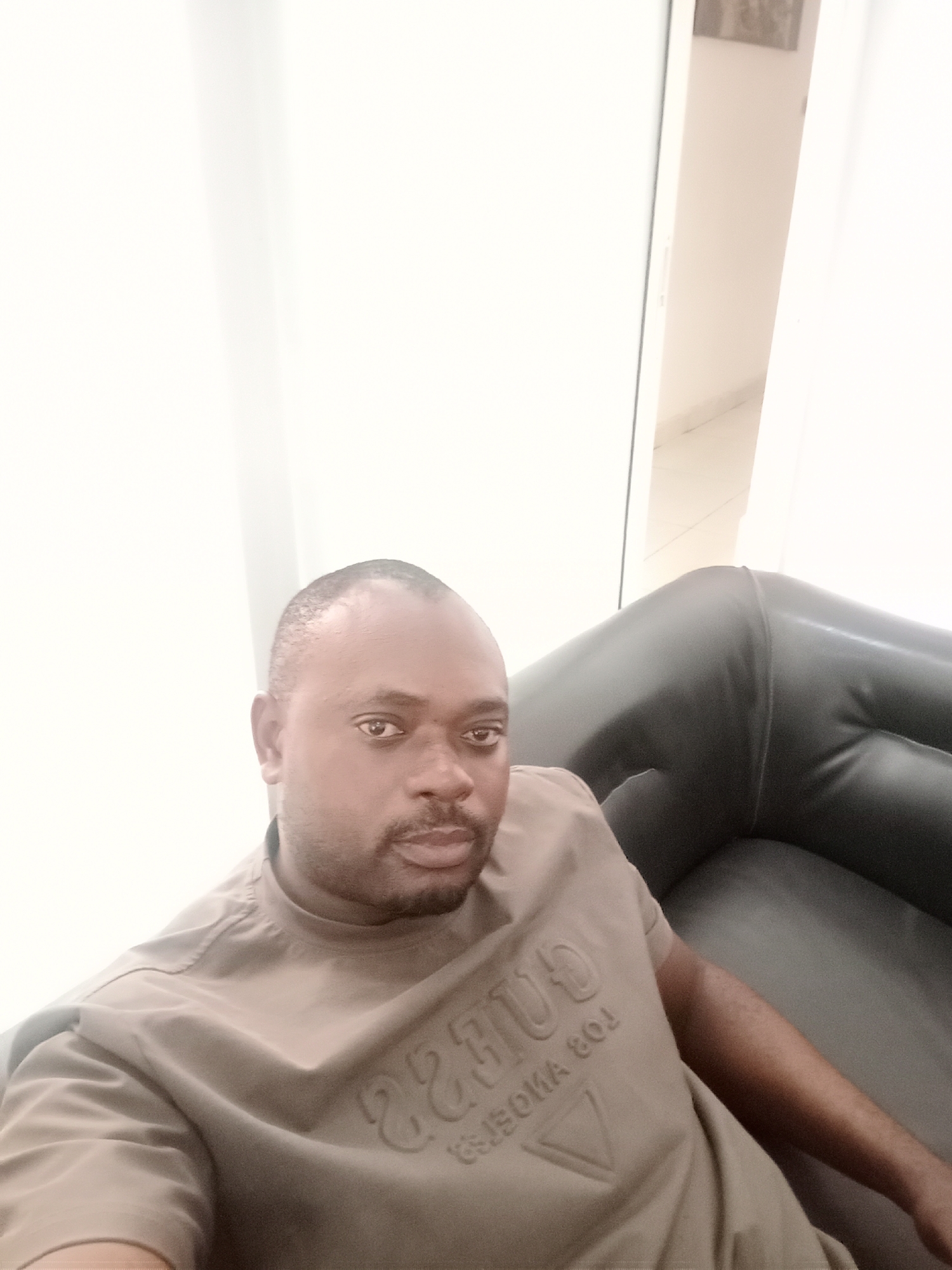 A medium shot depicts a man with short hair, a mustache, and a goatee, seated on a black leather couch in what appears to be an interior space. He is wearing a short-sleeved t-shirt with the "Guess" logo and lettering. The backdrop consists of a white curtain, and a partially visible open doorway. The location, Gwarinpa, Nigeria, is unverified from the image alone. No specific actions or interactions are evident.