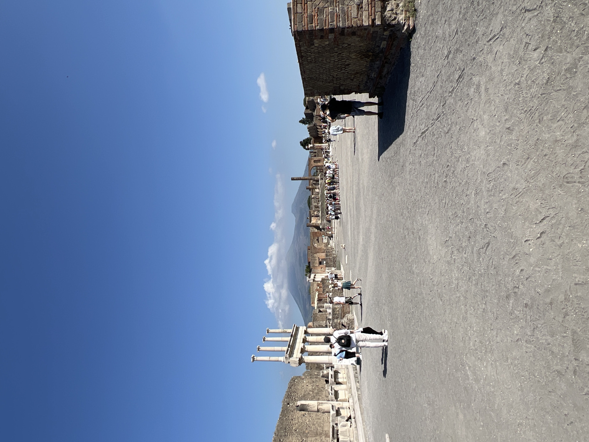 The image depicts a wide, open, paved area in the ancient Roman city of Pompeii, Italy. In the background, the imposing silhouette of Mount Vesuvius is visible, partially shrouded in clouds. The foreground is dominated by a textured gray ground, showing some unevenness and tire tracks.
To the left, there are ruins of ancient buildings, including a section of a stone wall and a prominent structure featuring several tall, fluted columns supporting an architrave. Several tourists are present, walking and observing the ruins. A group of four individuals stands near the columns, two facing away from the camera, one looking at a phone, and another woman walking. Further back, a larger group of tourists are gathered, appearing to be listening to a guide or observing a specific point of interest.
On the right side of the frame, a section of a brick wall with some stone accents is visible. A man wearing a dark t-shirt and shorts stands facing away from the camera, appearing to be taking a photo. Other scattered individuals can be seen in the middle ground. The sky is clear and a bright blue, with a few small, white clouds. The overall scene suggests a sunny day at a popular historical tourist destination.