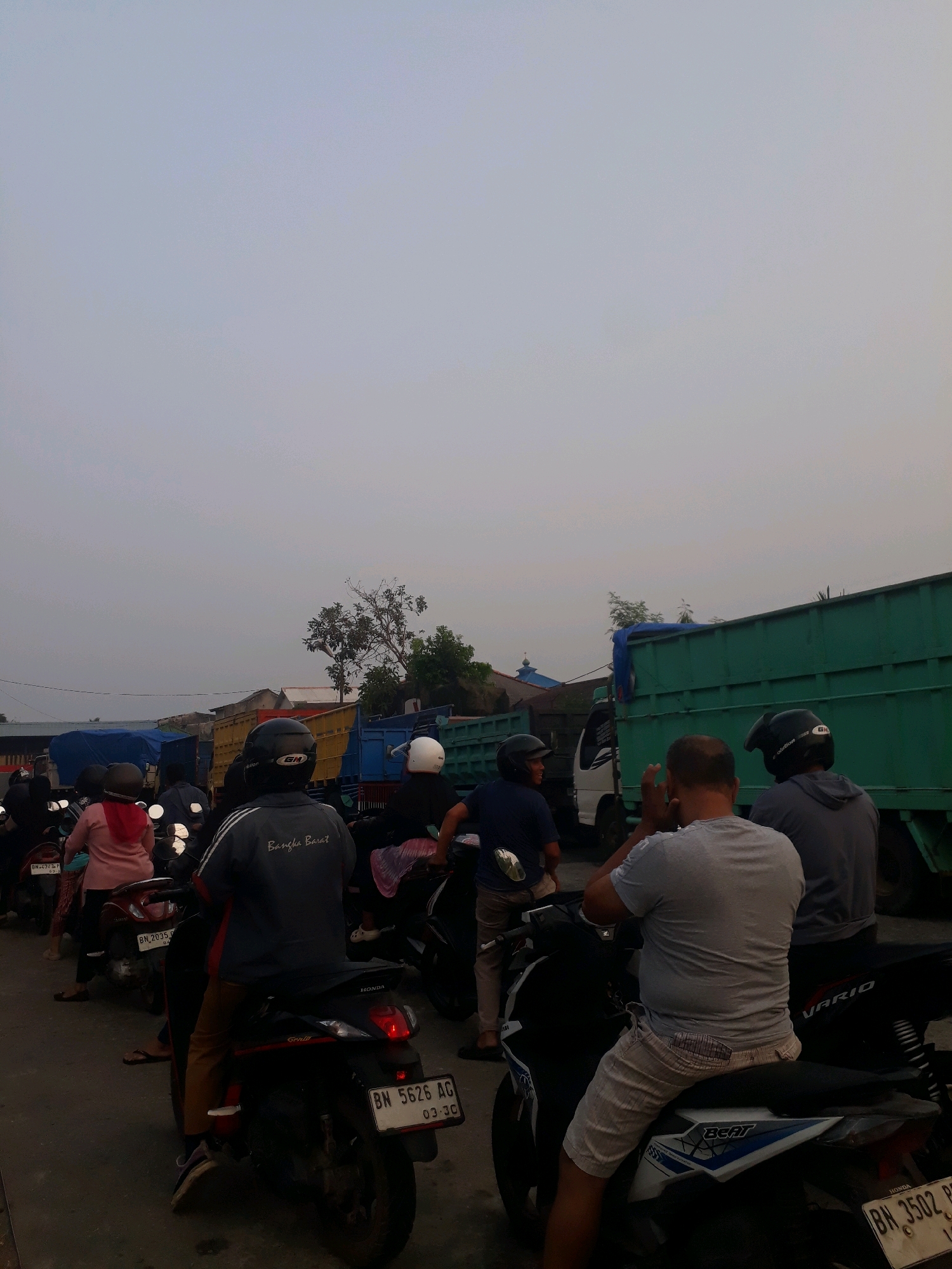 The image depicts a traffic jam in the city of Pangkalpinang, Indonesia. Several motorcycles and trucks are stationary on a paved road under a hazy, overcast sky. A few individuals are seated on their motorcycles, while others stand. One person in the foreground is wearing a grey t-shirt and light-colored shorts, sitting on a Honda Beat motorcycle. Another person in the middle ground wears a dark grey jacket with white stripes and "Bangka Barat" written on the back. The trucks are various colors, including green and yellow, with some carrying blue tarpaulin covers. The scene suggests a typical urban traffic situation in Pangkalpinang during daylight hours.