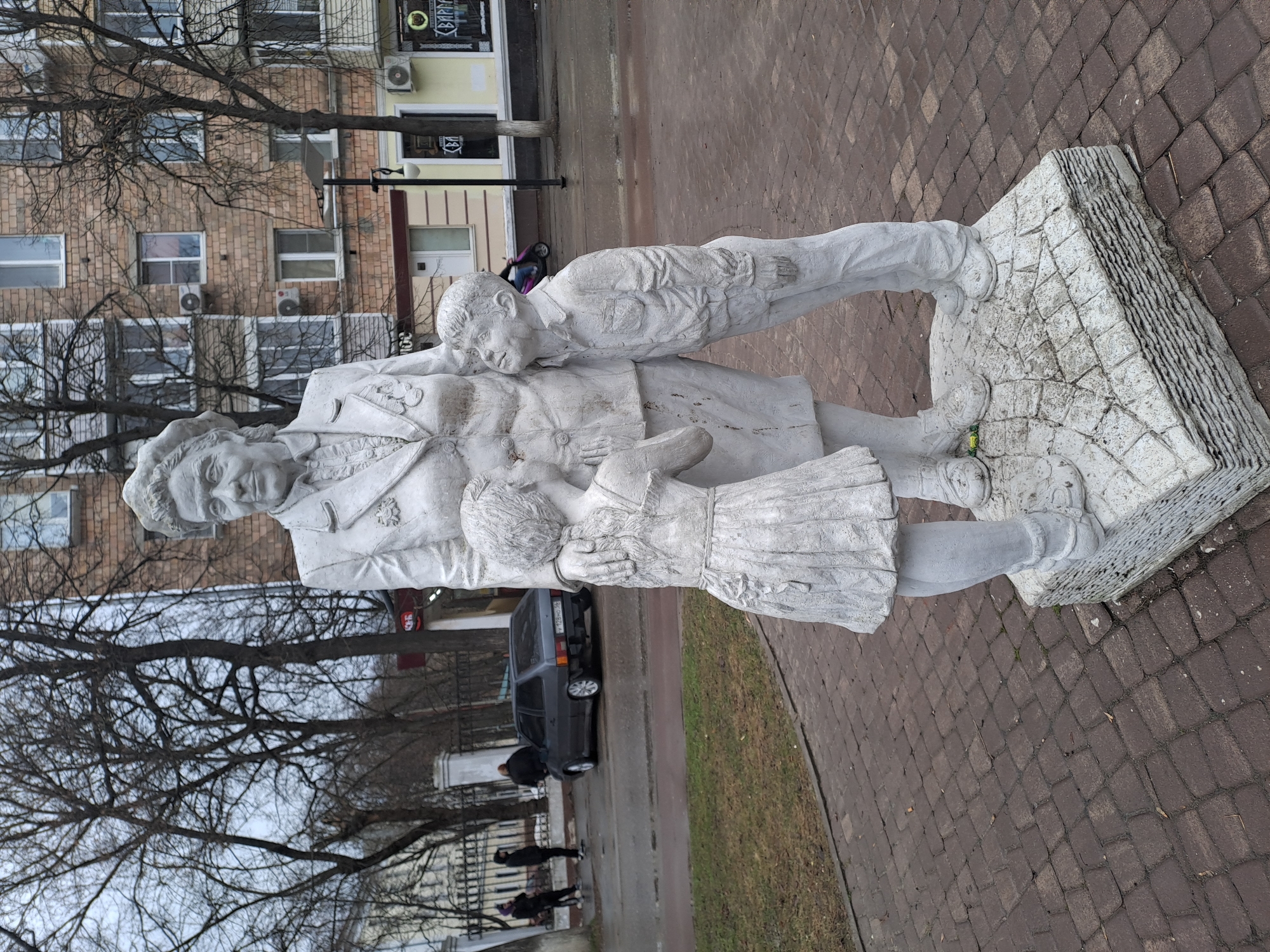 The image depicts a concrete monument in Elektrostal, Russia, featuring a woman with two children. The woman wears a jacket with a star on the lapel. A boy leans on her left shoulder and a girl stands in front of her, holding her hands. The monument rests on a textured, elevated stone base. The background includes a multistory brick building, bare trees, a street with parked cars, and pedestrians. The foreground shows a brick paved area. A building with a coffee shop is also visible in the background.