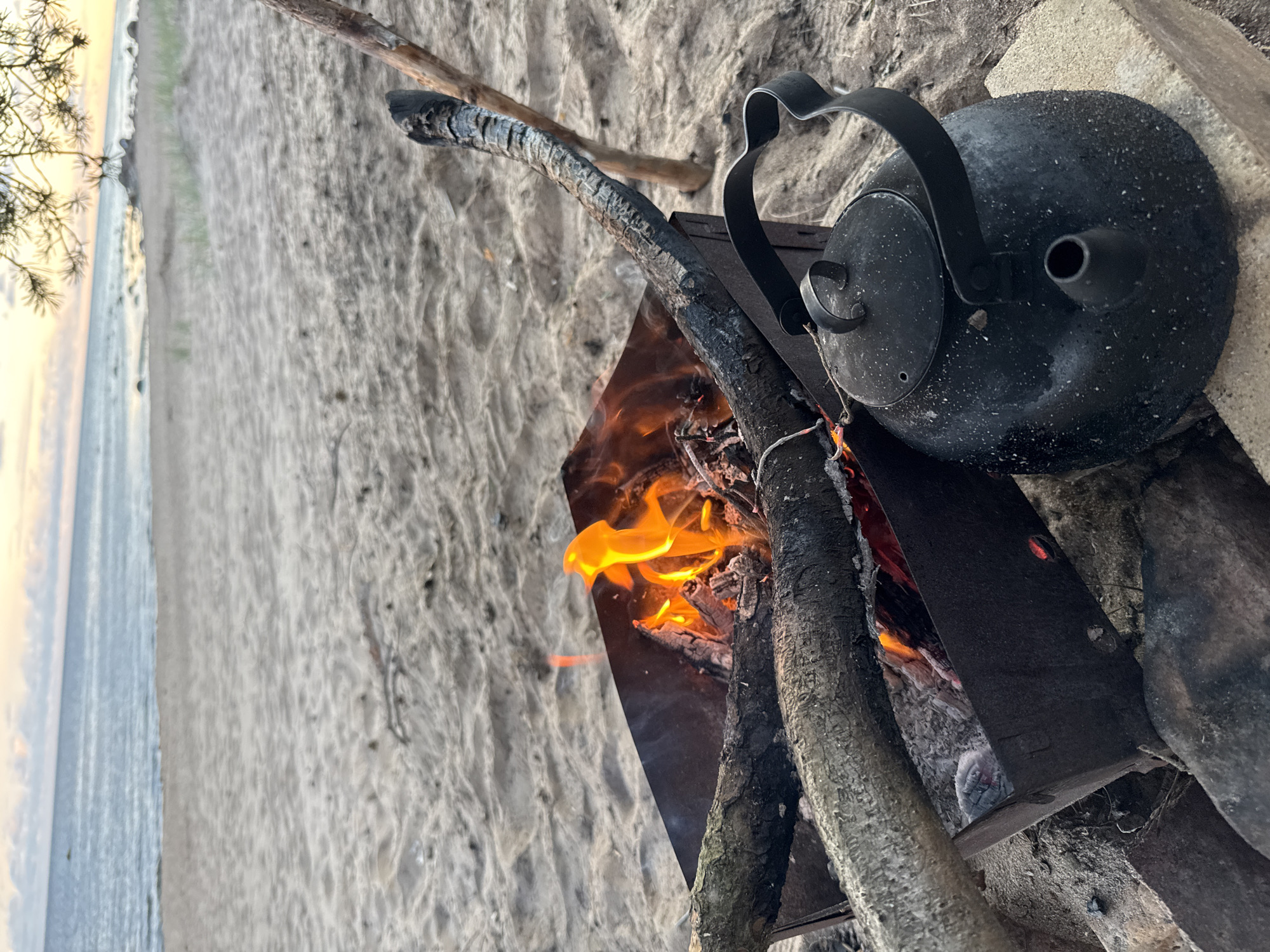 A campfire is burning in a metal brazier on a sandy beach, with logs and a black kettle placed nearby. The sea is visible in the background under a pale sky. The scene appears to be in Shepelevo, Russia, suggesting a coastal outdoor setting.