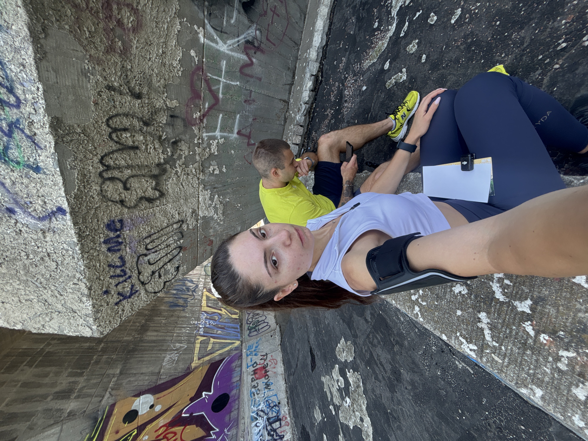 A young woman with brown hair in a ponytail, wearing a white tank top and dark leggings, is seated on a concrete ledge. She is holding a phone in her right hand, and her left arm is adorned with an armband. Beside her, a man wearing a yellow t-shirt and dark shorts is also seated, looking at his phone. The background features a concrete structure covered in graffiti. The location is the area beneath the Asparuhov most bridge in Varna, Bulgaria. The ground is dark and appears wet in some areas, with scattered debris. The lighting suggests it is daytime.