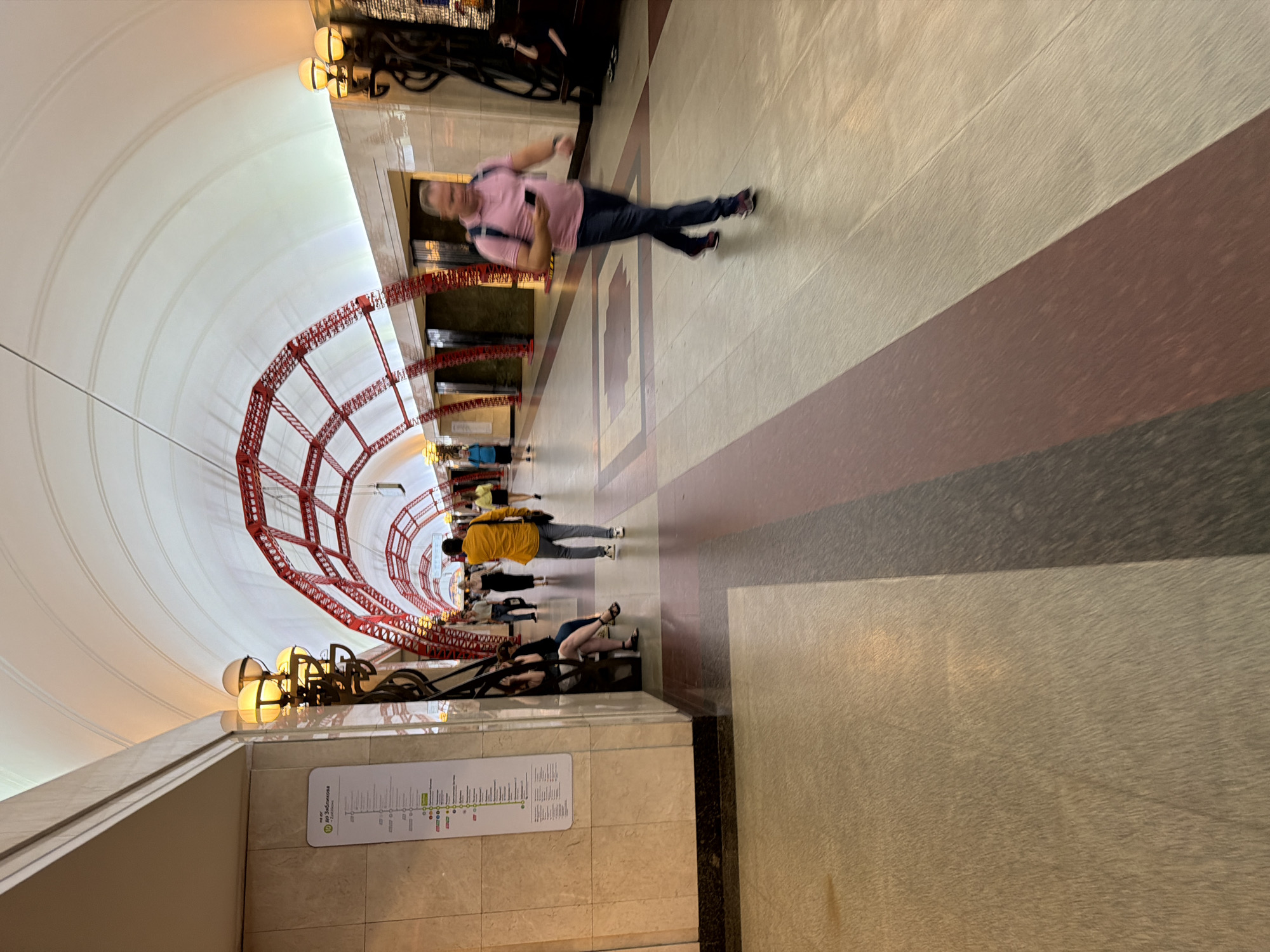 The image shows a wide, arched subway tunnel with ornate red metal structures supporting the ceiling. The walls are tiled and illuminated by golden lamps. On the left, a vertical subway map is displayed. Several people are walking through the tunnel. In the foreground, a man in a pink shirt and dark pants is walking away from the camera, looking at his phone. Further down the tunnel, a person in a yellow shirt is walking towards the camera. Other individuals are visible in the background, some sitting on a bench. The floor is a patterned tile with bands of light brown, reddish-brown, and dark gray. The overall impression is of a grand and well-maintained public transport space.