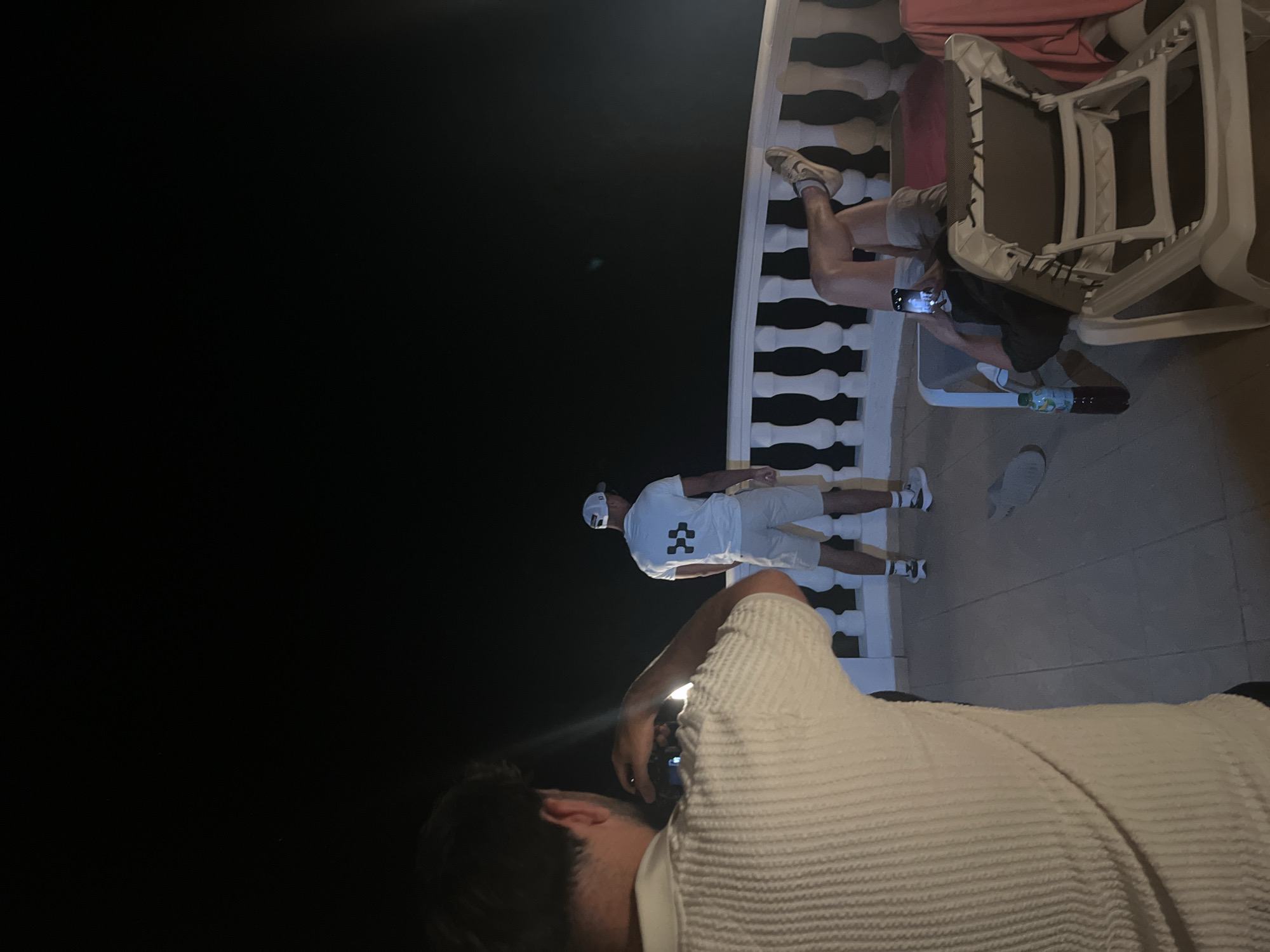 Here is a description of the image:
This is a night scene, apparently taken at a balcony. From the angle, it appears someone is taking a picture of another person standing on the balcony, near a white decorative balustrade. The person being photographed is wearing all white clothes including a cap. There are other people nearby sitting down. A white plastic chair is also visible. The location is likely in or near el Poble Nou de Benitatxell / Benitachell, Spain, although this is based on the image's source and not visual details in the image itself.