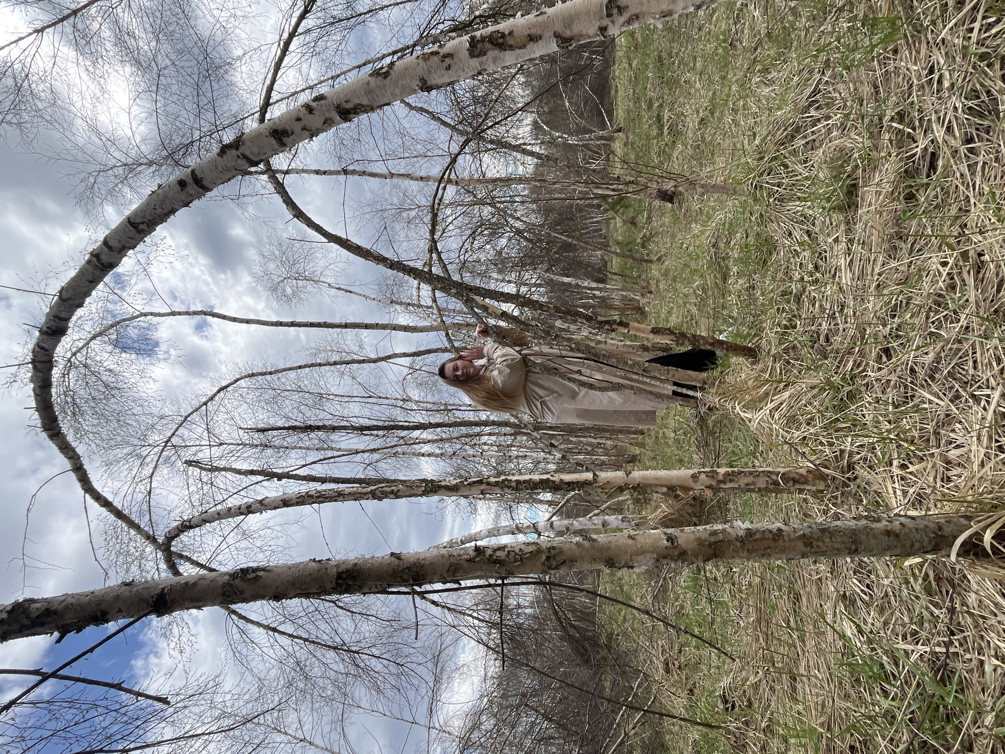A woman stands in a birch tree forest in Razdolie, Russia. She wears a tan trench coat. The ground is covered in dry grass and dead leaves. The sky is overcast with a mix of blue and grey clouds. The trees are thin and mostly bare of leaves.