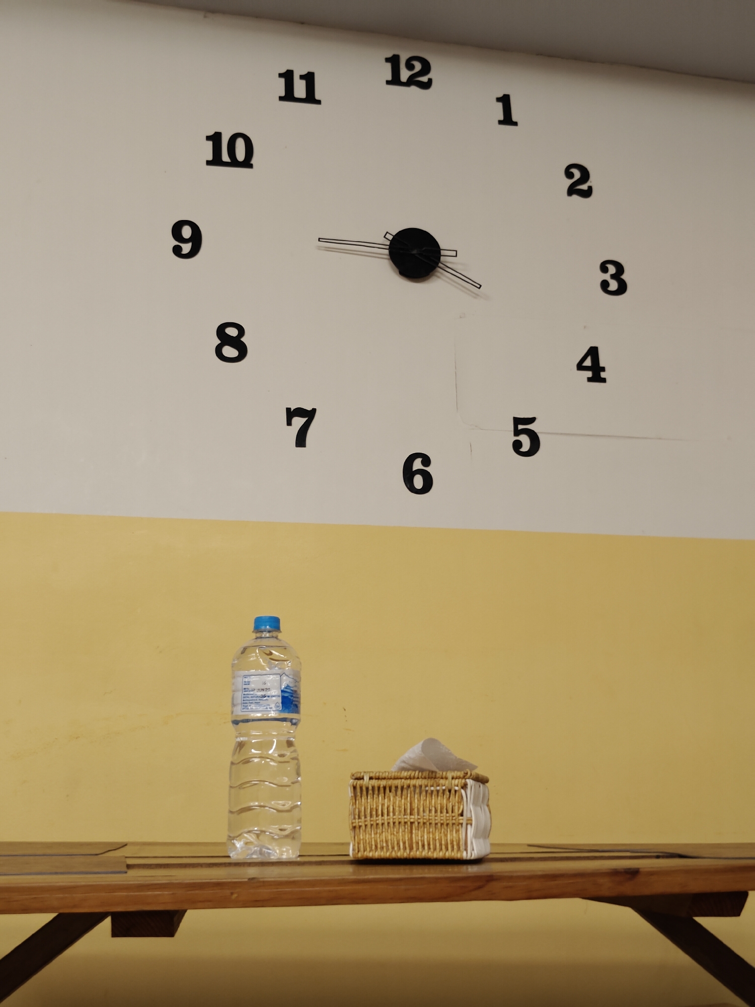 A modern clock with large, black numerals on a white wall is the dominant subject. The clock's hands are visible, indicating a time slightly past 2:15. Below the clock, a wooden table is set against a wall painted with a distinct yellow stripe across its lower half. On the table, a clear plastic bottle of water with a blue cap stands next to a small, woven basket filled with white tissues. The scene appears to be indoors, possibly in a casual waiting area or common space. The lighting is neutral, suggesting an indoor setting without strong natural light sources. No people are present in the image. The water bottle has a label with some text, including "JUN 20" which could indicate an expiration date. The overall impression is one of simplicity and practicality, with common household or public space items arranged neatly.
