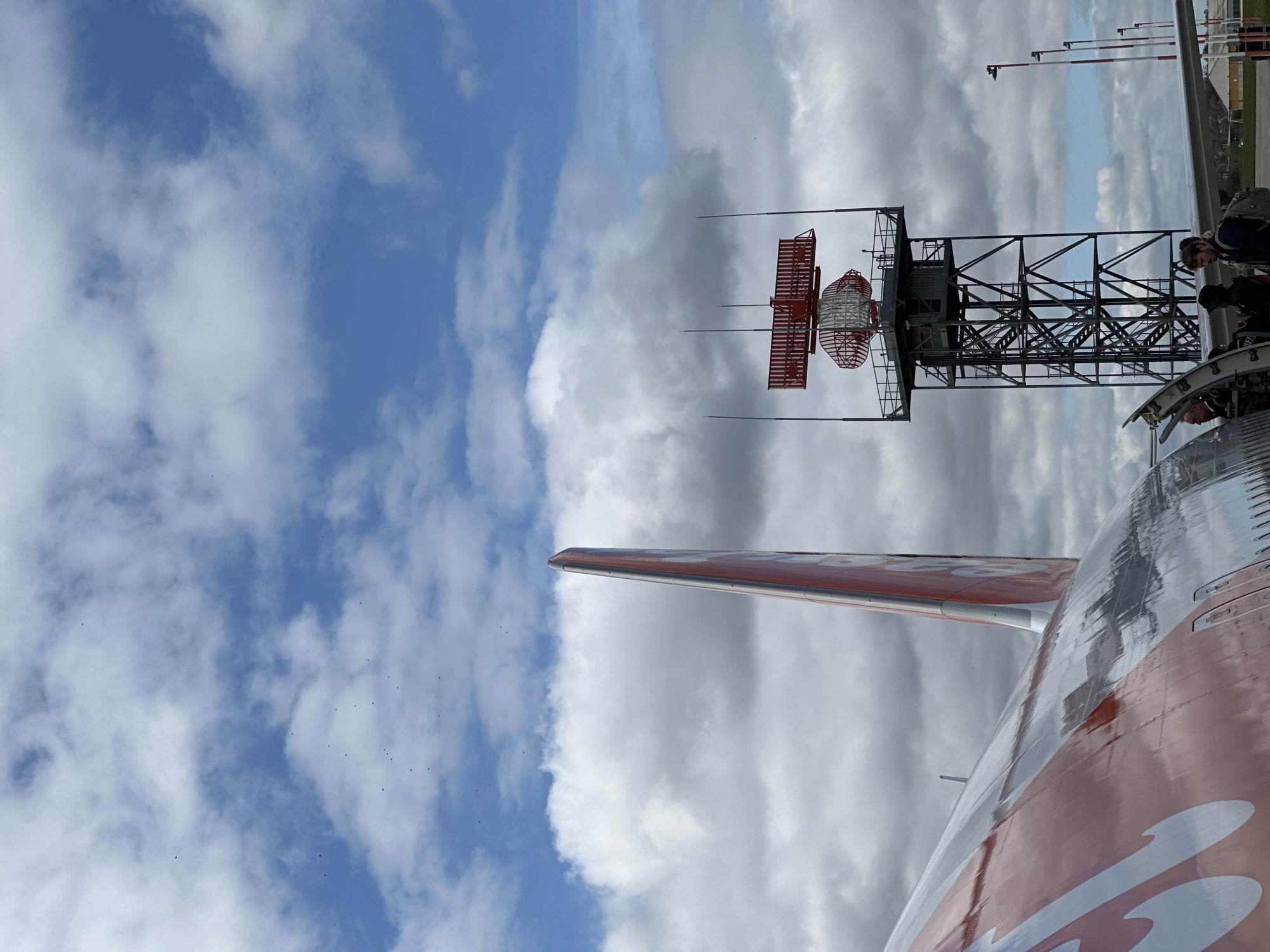 The image shows a partial view of an orange and white airplane with the tail fin visible against a backdrop of a cloudy sky. The location is Rochford, United Kingdom as evidenced by the Rochford Air Traffic Control Radar tower in the background. There is a person near the edge of the plane. Several birds are flying in the sky.