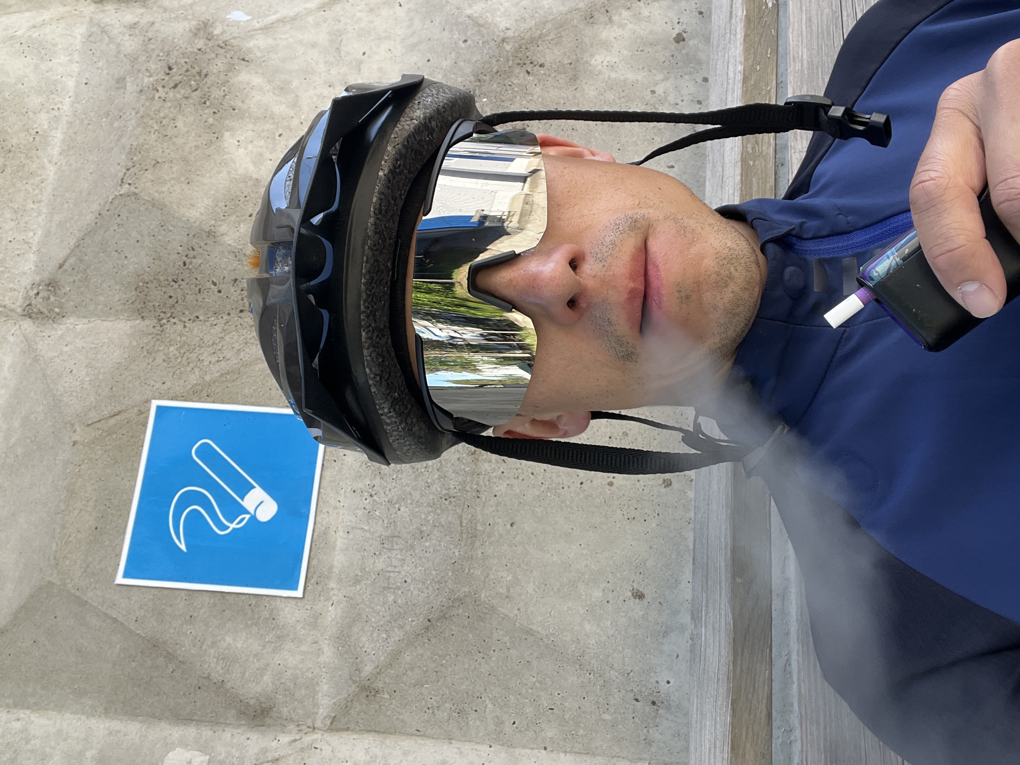 A man with a helmet and sunglasses is seated in Chaikovsky, Russia. He wears a blue jacket. The man is holding a cigarette and exhaling smoke. A blue sign on the ground depicts a cigarette with smoke. The location is outdoors on a gray surface.