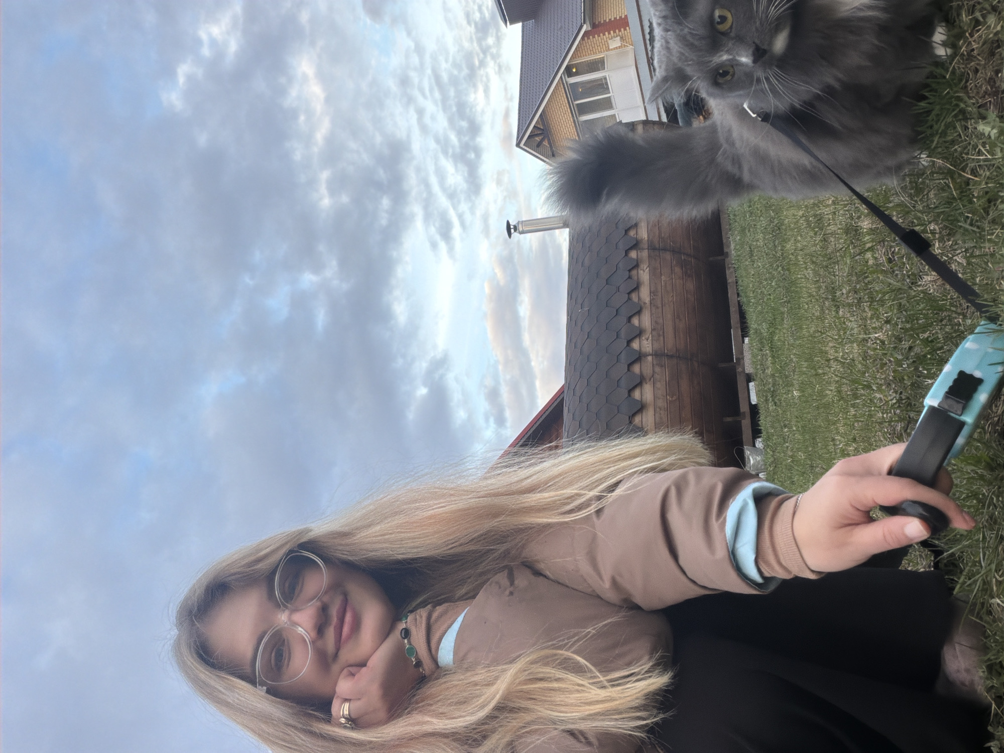 The image shows a woman with long blonde hair and glasses, wearing a brown jacket, holding a leash handle, with a gray cat on a leash in the background. Behind the cat is a wooden structure with a dark-colored roof, and further back is a two-story house with light-colored brick on the upper level. The sky is cloudy with some visible blue patches. Based on the available visual cues, the location is in the city of Большая Шильна, Russia.