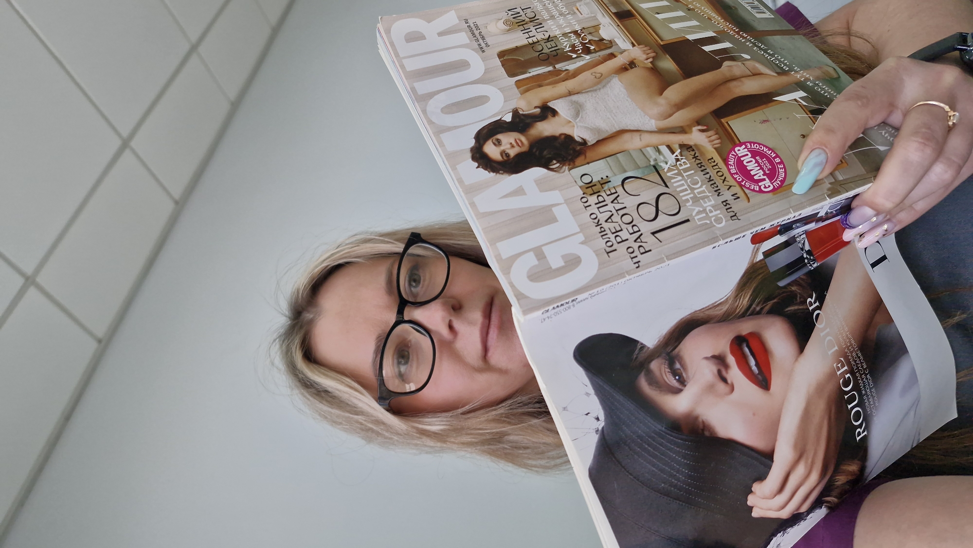 Here's a detailed description of the image:
A woman with shoulder-length blonde hair and glasses is holding two open copies of the Russian magazine "Glamour." The magazine in front features a woman with red lipstick from Rouge Dior. The cover states that there are 182 beauty products, which were the best beauty products of 2021. The magazine at the top features a brunette woman. The woman's nails are painted in different patterns, and she has a ring on her finger.
The location may be Otchor, Russia.