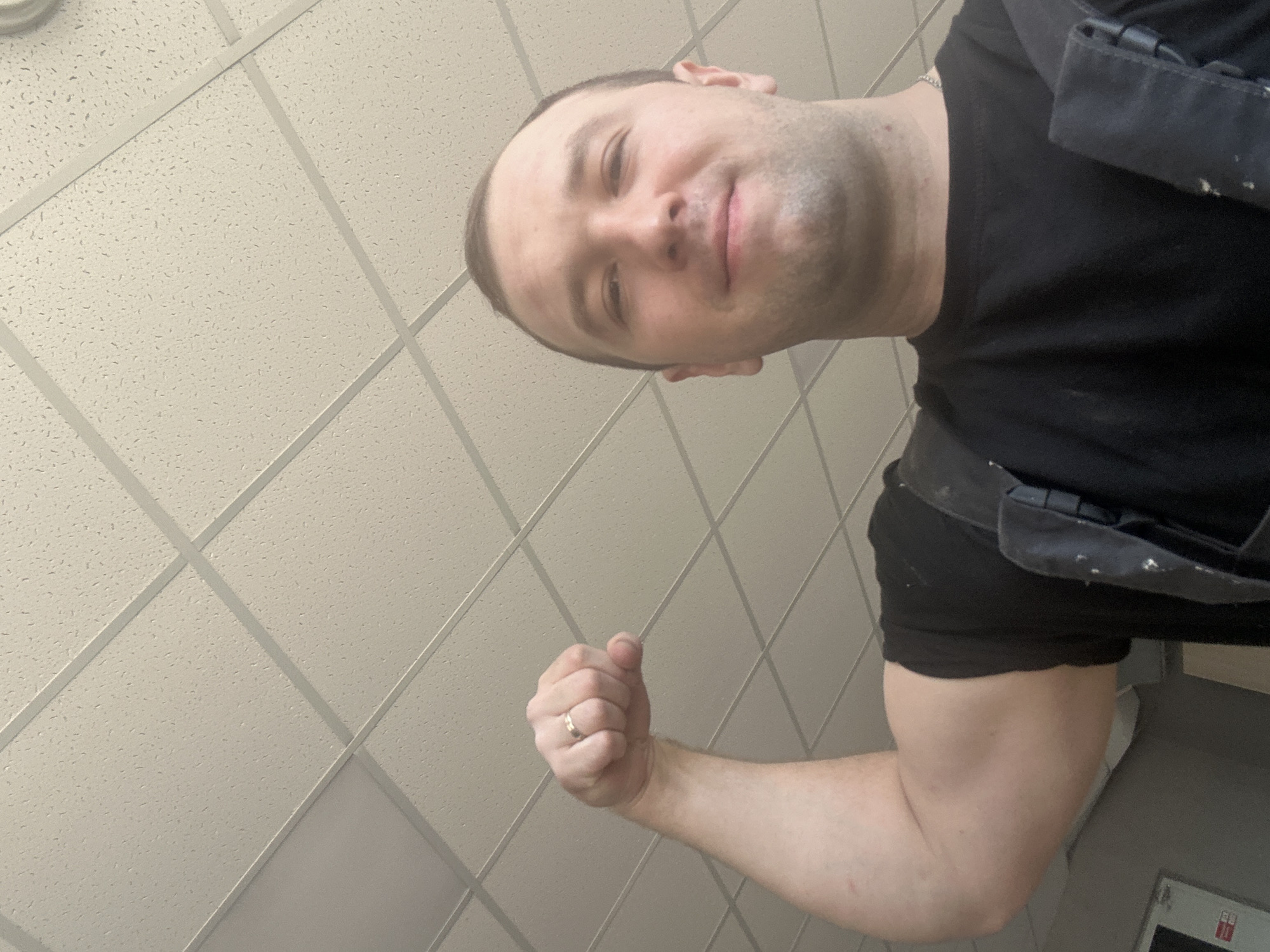 A low-angle selfie of a light-skinned man wearing a black t-shirt and dark work pants. He is flexing his right arm and making a fist, on which a gold ring is visible. The background is a drop ceiling with square, textured tiles. No evidence exists in the image that would indicate that the location is in the city of Noviy, Russia.