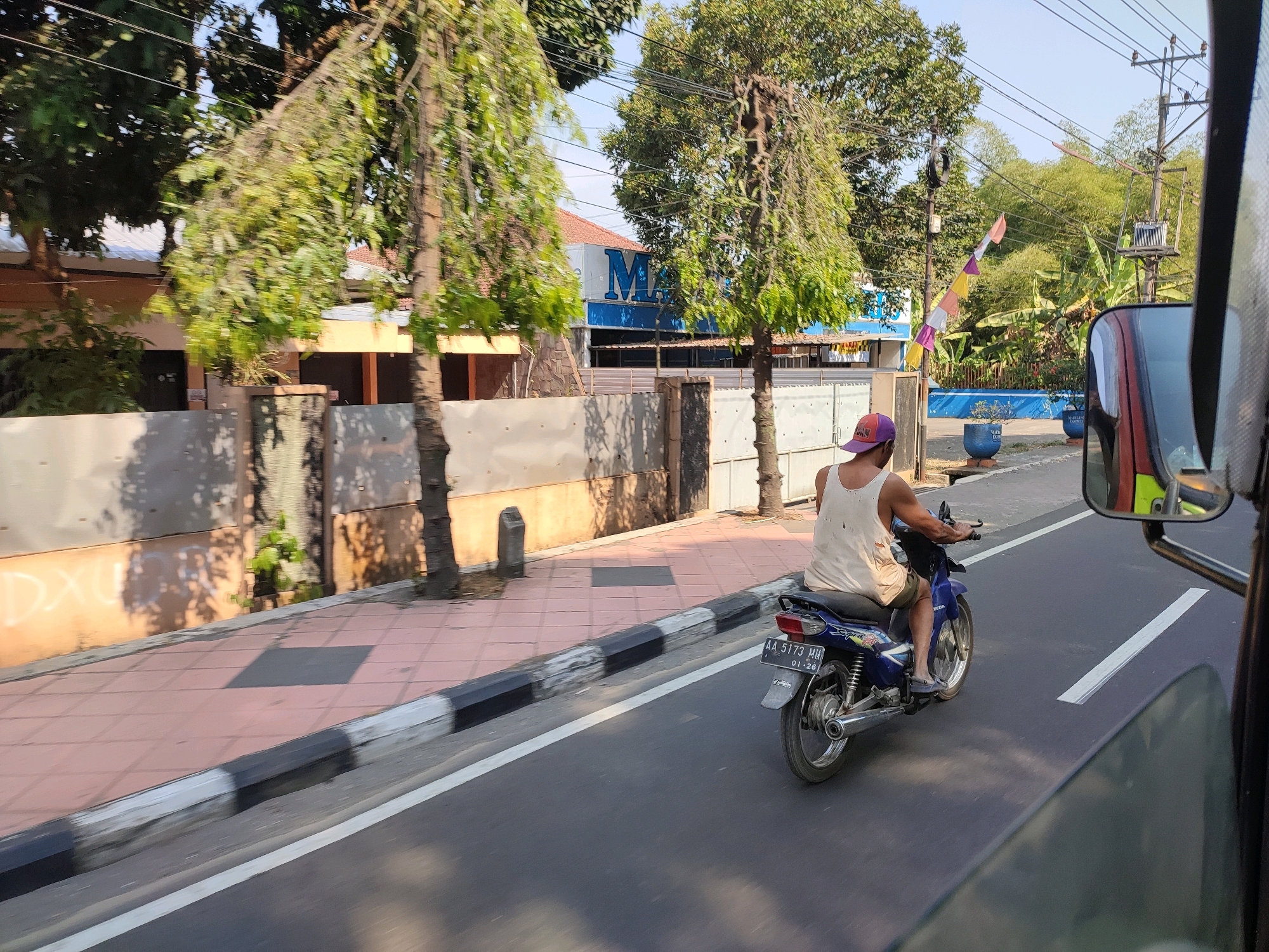 The image shows a view from a vehicle, likely a car, looking out at a street scene in Magelang, Indonesia. A person wearing a purple cap and a white tank top is riding a blue motorcycle on the right side of the road, moving away from the viewer. The motorcycle has a license plate reading "AA 5173 MH 01-26". On the left side of the road, there is a sidewalk paved with pink tiles and a raised curb with black and white stripes. Behind the sidewalk is a wall with some graffiti and a building with an orange facade. Further down the street, there are trees, power lines, and what appears to be a building with a blue sign that has a white "M" on it. In the background, there are more trees and vegetation. The sun is shining, casting shadows on the sidewalk and road. A side mirror of the vehicle is visible in the foreground on the right.