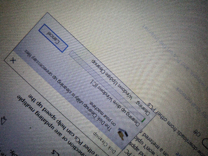 The image shows a laptop screen displaying a Disk Cleanup utility pop-up window in progress. The utility is "clearing up unnecessary files on your machine." The progress bar shows that "Cleaning up drive Windows (C:)" is completed, and "Cleaning Windows Update Cleanup" is in progress. The Cancel button is visible in the top right corner of the pop-up window. The laptop is located in Spokane, Washington, USA. The coordinates provided (latitude: 47.655, longitude: -117.379) correspond to a residential area in Spokane. The image captures a close-up of the laptop screen, focusing on the Disk Cleanup utility and its progress bar. The background shows a portion of the laptop screen, with blurred text. The image provides a glimpse into a user's computer activity and indicates that they are performing a Disk Cleanup task.