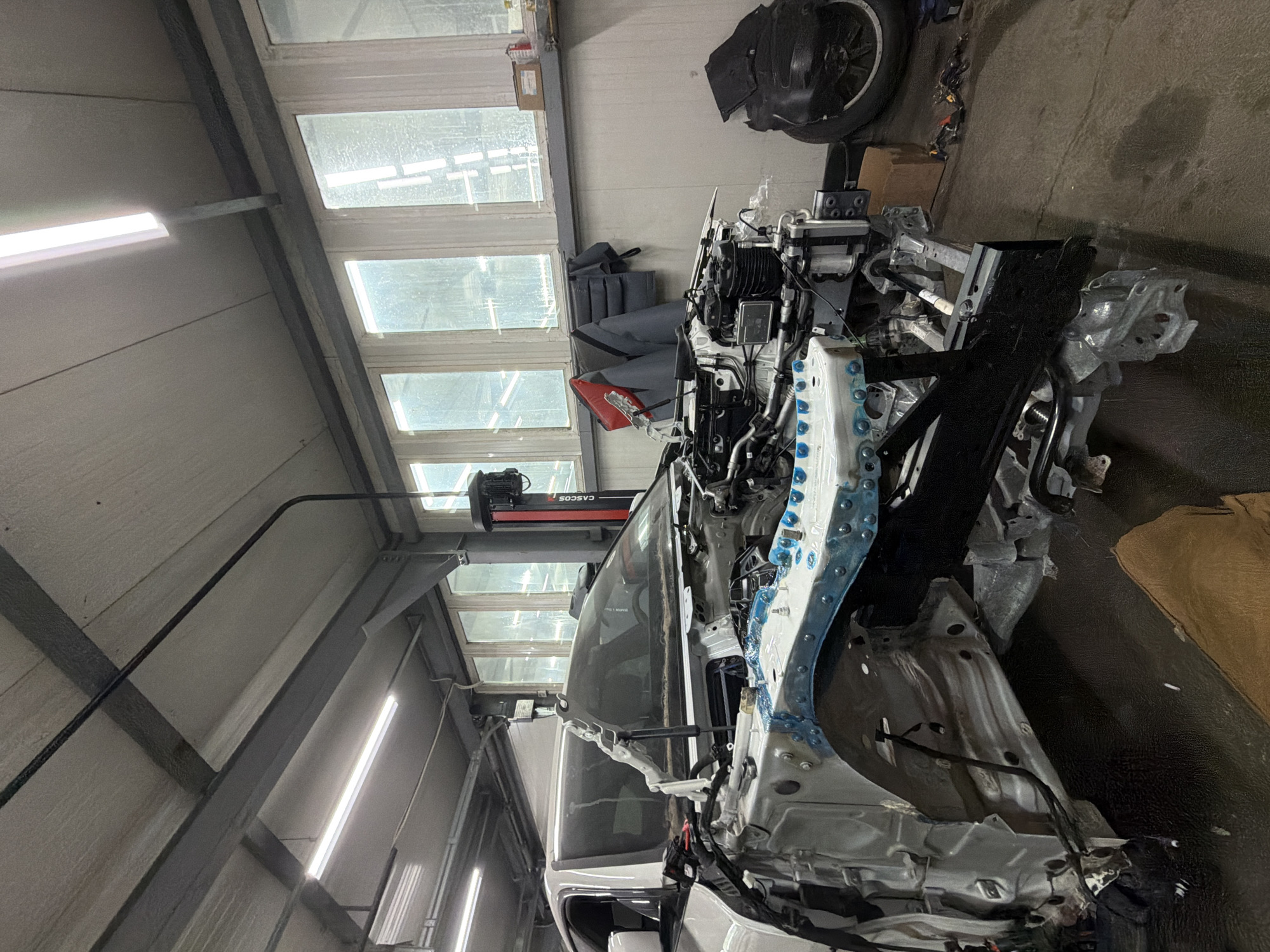 The image shows a disassembled white car in what appears to be a repair shop. The front section of the car is exposed, revealing the engine and various mechanical components. The car is white and appears to be a modern passenger vehicle. The workshop is well-lit with fluorescent lights on the ceiling. There are large windows in the background, showing an overcast sky. A red hydraulic lift is visible on the right side of the car. Several items are placed around the car, including a cardboard box, a black tire, and some tools. The floor of the workshop is dark and appears to be concrete or asphalt. The image does not provide any direct clues to identify the specific location in Burgas, Bulgaria.