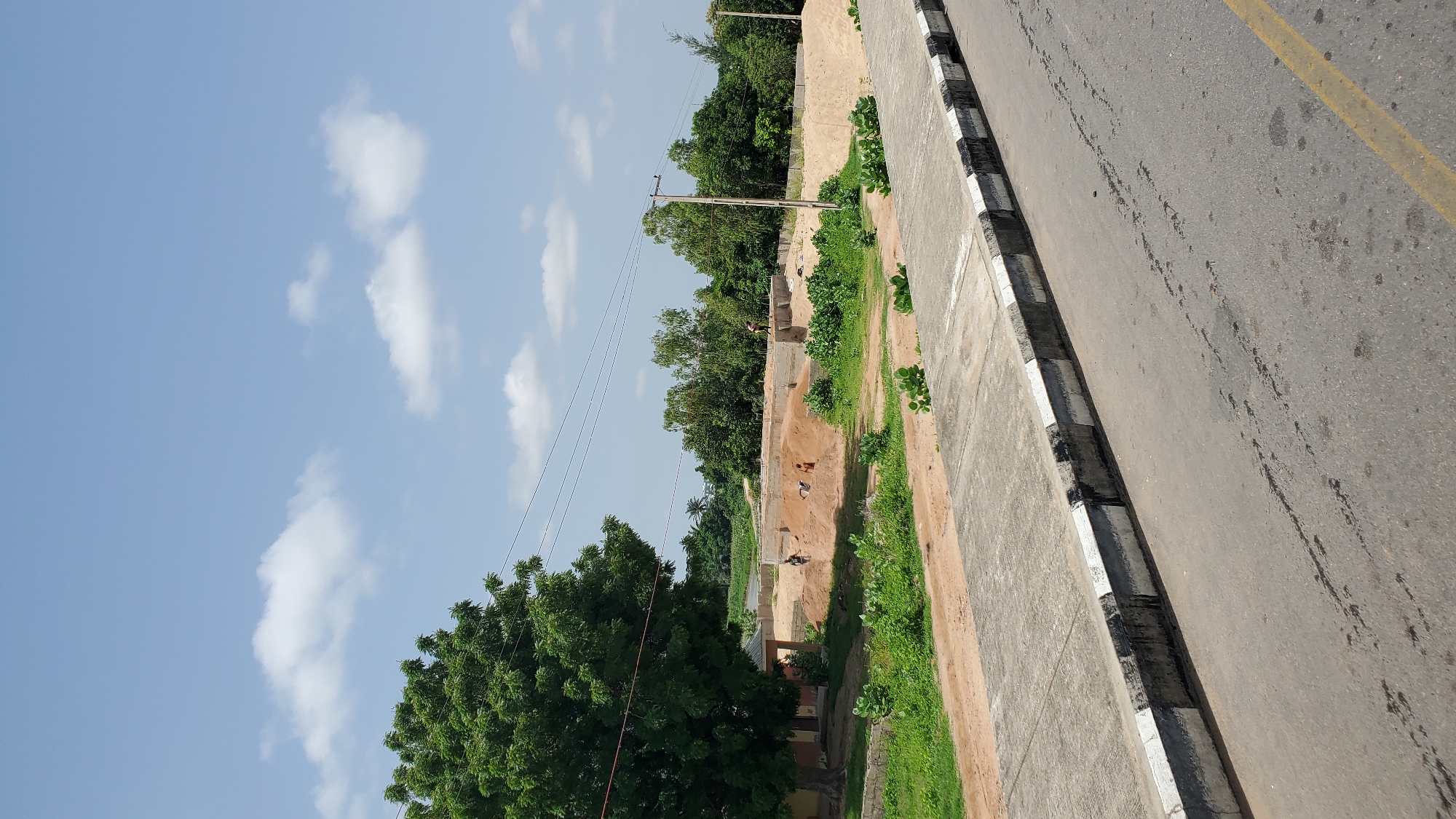 In Jalingo, Nigeria, a construction site is visible in the mid-ground, featuring several individuals engaged in labor on the foundation of a building. Piles of sand and concrete structures indicate ongoing construction activities. The foreground of the image displays a paved road with distinct black and white painted curbs, suggesting an urban or semi-urban setting. A large, leafy tree stands to the left of the construction site, providing shade and a touch of greenery. The background is characterized by a dense arrangement of trees, indicative of a natural or semi-rural environment. The sky above is clear with a few scattered white clouds, suggesting a daytime scene.