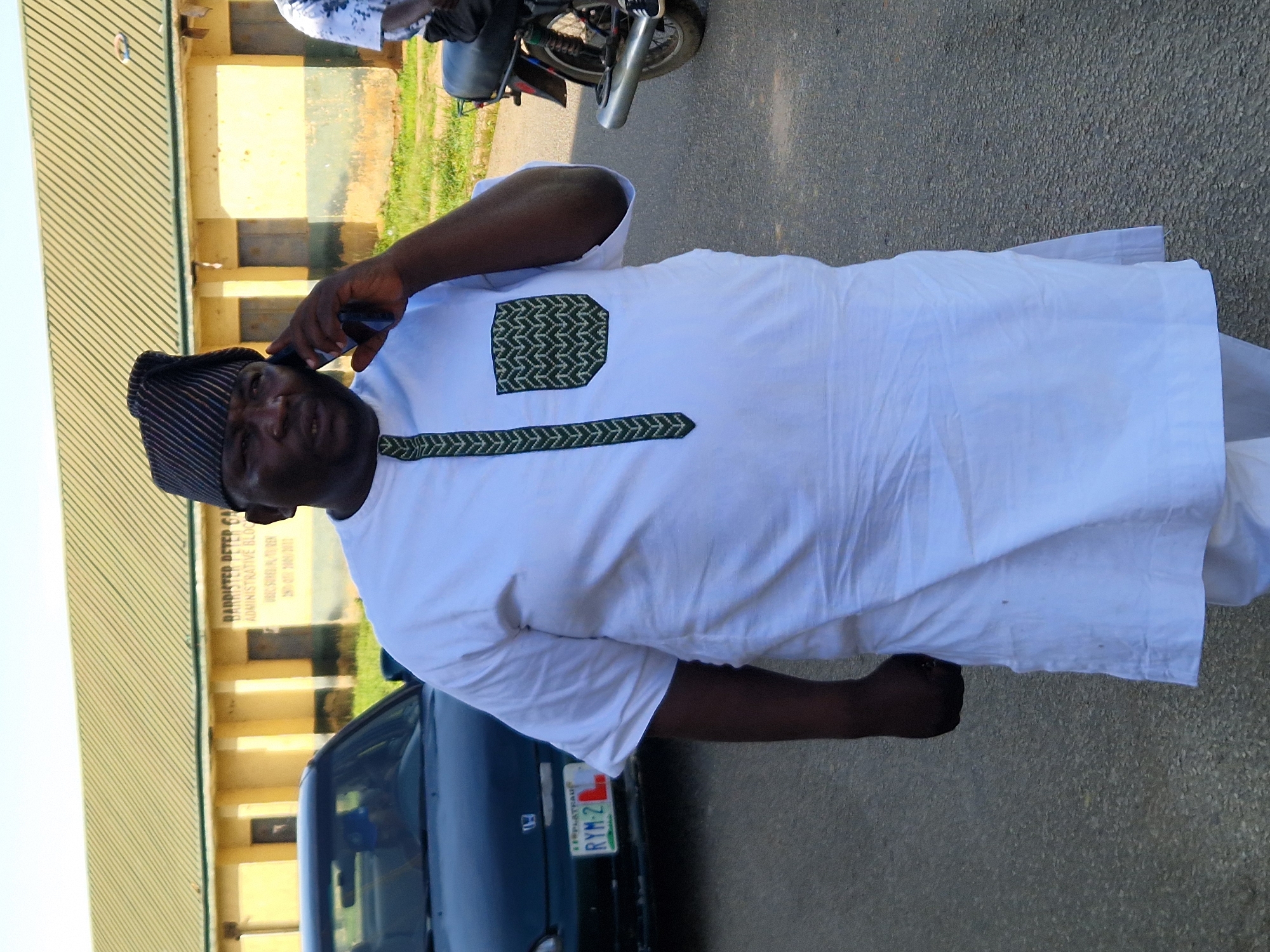 A full-body, eye-level shot shows a Black man standing outdoors, wearing a white kaftan and a black and white striped cap. He is holding a dark-colored cell phone to his right ear with his right hand, and his left hand is clenched in a fist near his stomach. The man's kaftan has a green and black striped pattern down the front and a patterned pocket on the right side of his chest. The man appears to be in his late 40s or 50s, with a dark complexion and a bald head under the cap.
Behind the man, a dark blue car is parked, with its front bumper and license plate visible. The license plate reads "RYM-2 L", with "PLATEAU" above it. To the right of the man, a motorcycle is parked, with its rear wheel and part of its exhaust pipe showing. In the background, a yellow building with a green corrugated metal roof is visible, with signs on the wall. One sign reads "BARRISTER PETER GA", and another smaller sign below it reads "ADMINISTRATIVE BLOC". The ground is paved with asphalt, and there is some green grass and foliage visible in the background. The sky is a pale blue. The scene appears to be in an urban or semi-urban environment. Based on the license plate, the location is likely the city of Kabong, Nigeria.