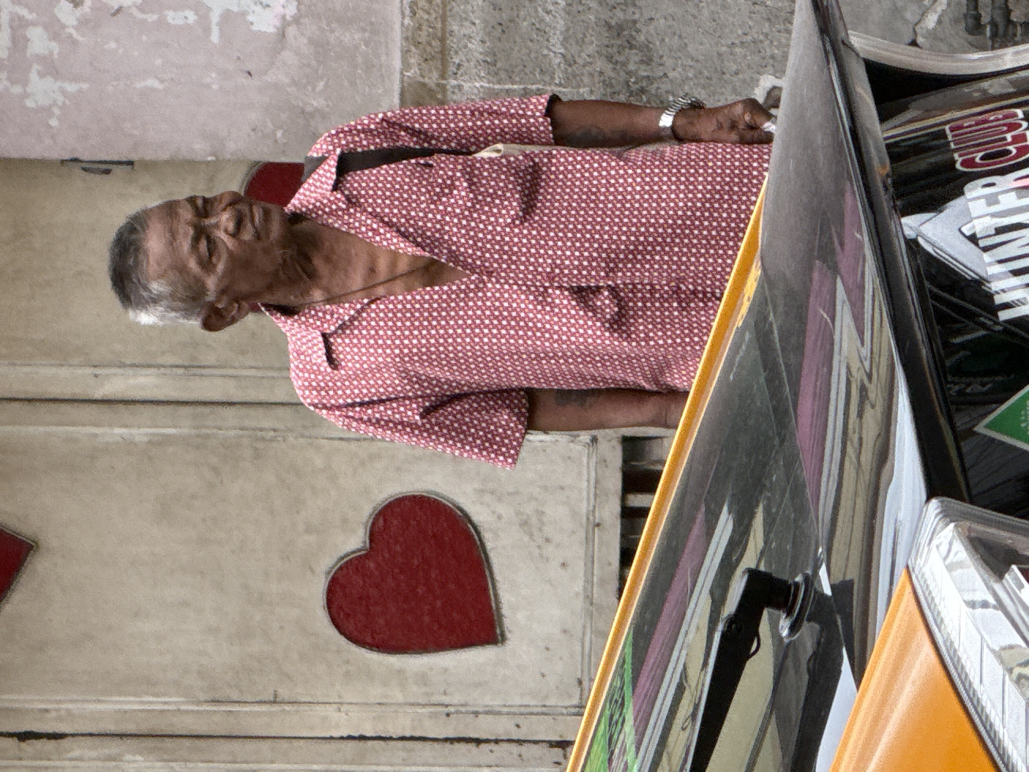 An elderly man with graying hair and weathered skin stands in front of a weathered wall with two red heart decals. He is wearing a short-sleeved, collared shirt with a small white geometric pattern on a red background and a necklace. A dark strap is visible over his right shoulder, suggesting a bag. His right hand, adorned with a wristwatch, rests on his hip. In the foreground, the edge of a yellow vehicle with chrome accents is visible, along with a dark, reflective surface that appears to be part of a larger vehicle, possibly a tuk-tuk. The overall scene suggests a street environment in Bangkok, Thailand, likely in a bustling urban area given the presence of the vehicle. The lighting is overcast, casting soft shadows.