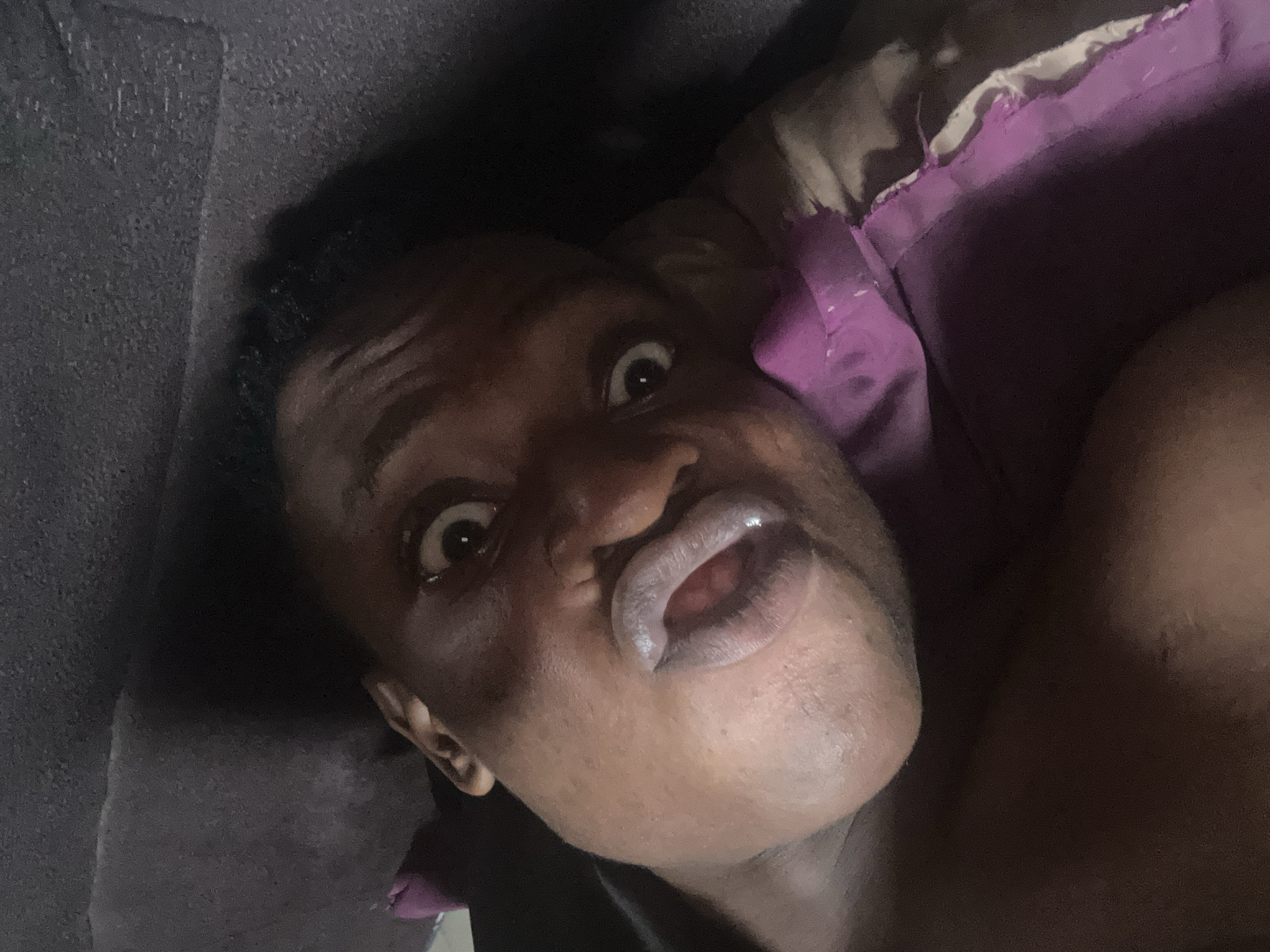 A close-up, overhead shot shows a person lying down, with their face taking up most of the frame. Their eyes are wide open, and their mouth is agape, with their lips slightly parted. The person's skin tone is dark, and their hair is short and curly. They are lying on a dark-colored surface, possibly a bed or couch, with a purple pillow visible on the right side of the frame. The lighting is dim, casting shadows on the person's face and the surrounding environment. The background is dark and out of focus, suggesting an indoor setting. The image appears to be a selfie, taken by the person themselves. No specific location in Ikorodu, Nigeria can be determined from the image.