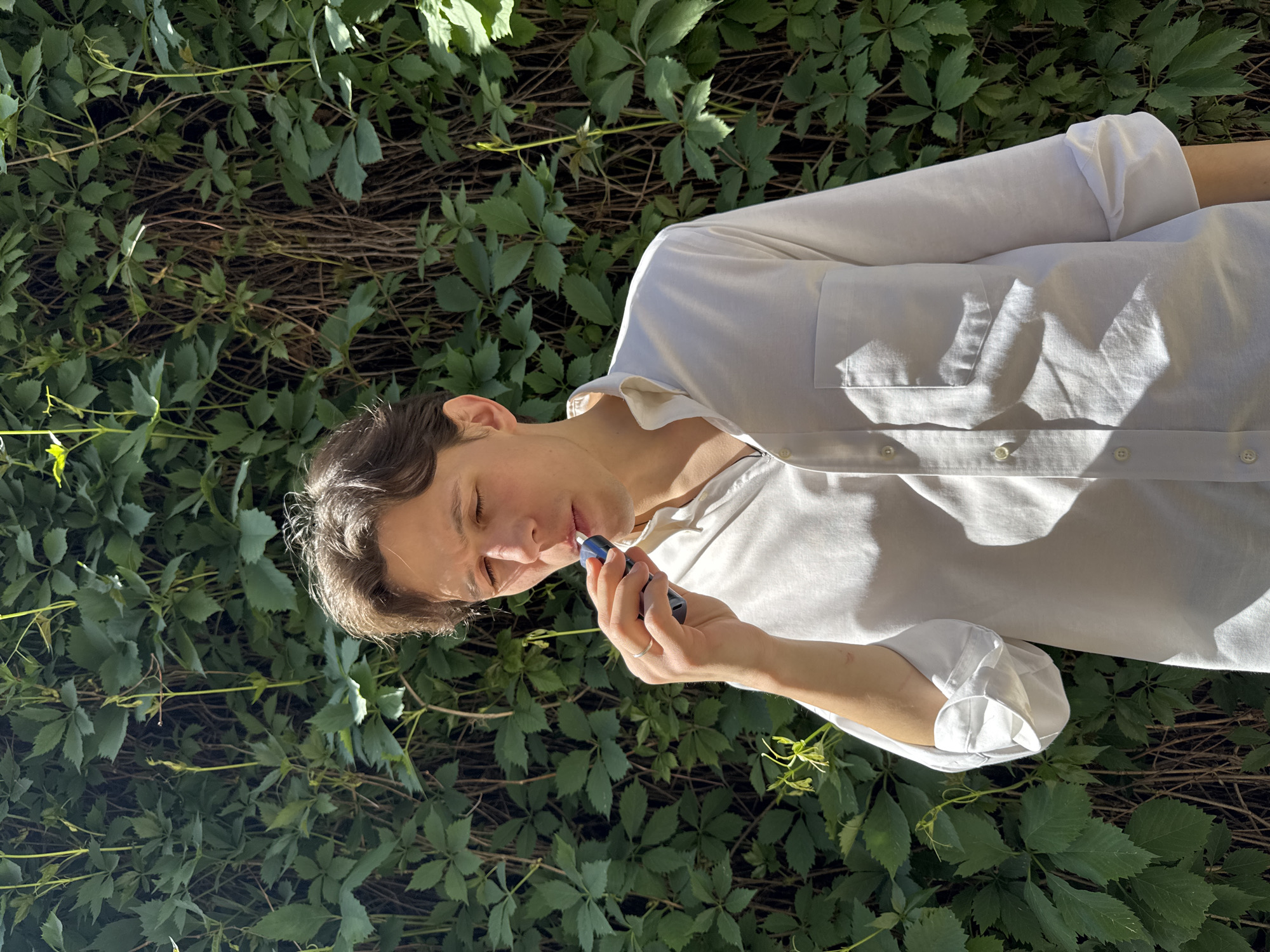 A fair-skinned person is applying lip balm. The person is wearing a white collared shirt and is positioned in front of a wall covered in green foliage. The location could be Moscow, Russia, but this is not certain.