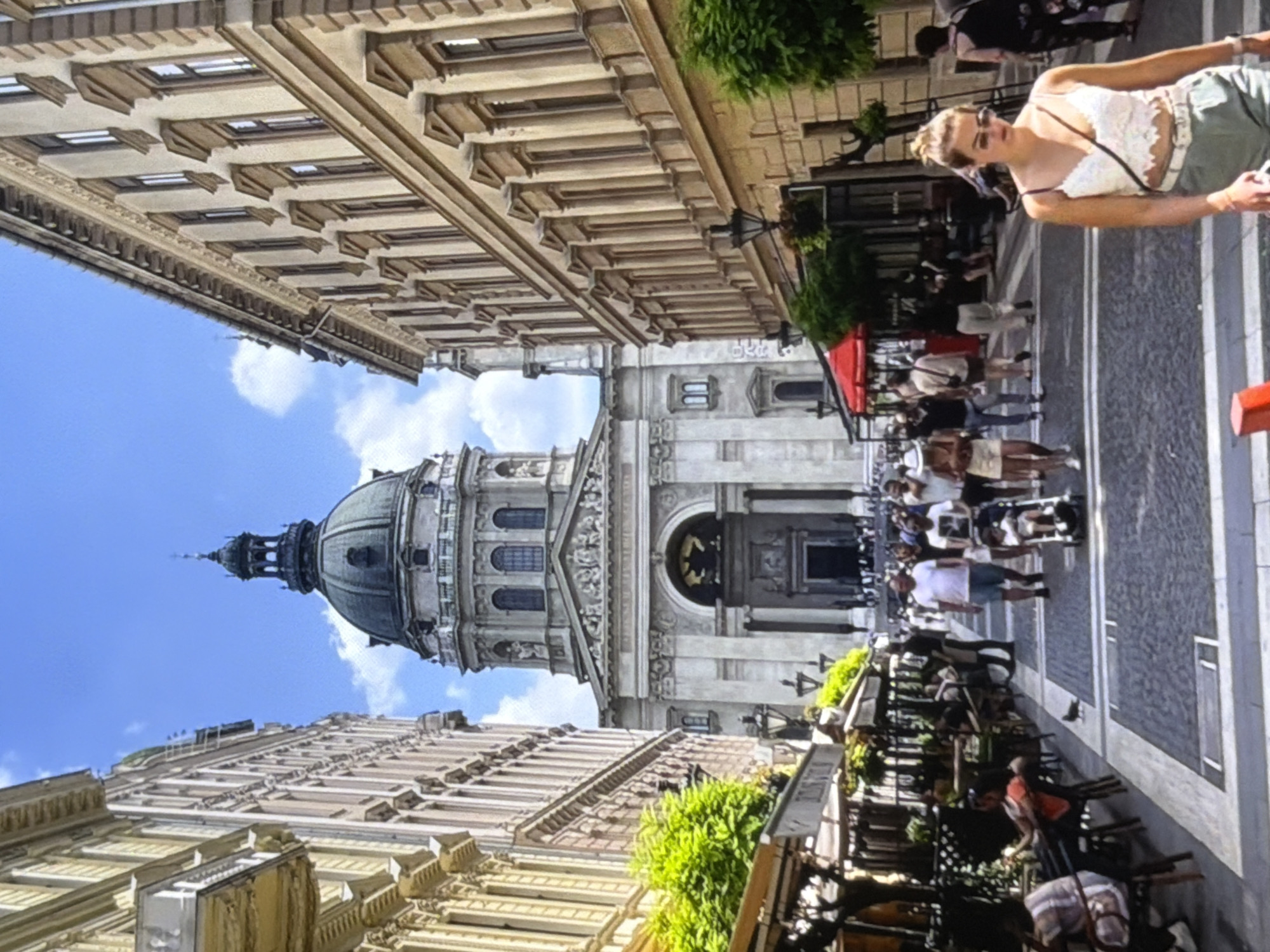 In a European city, St. Stephen's Basilica, a grand neoclassical church with a large dome, dominates the background of a busy pedestrian street. The street is lined with multi-story buildings featuring classical architectural elements, such as ornate balconies and rows of windows. On the right side, cafes with outdoor seating and umbrellas are set up along the sidewalk, with people dining and socializing. A diverse group of pedestrians walks along the street, some pushing strollers, others conversing. A pigeon is visible on the cobblestone street in the foreground. A young woman in a white lace top and green shorts walks towards the camera, wearing sunglasses. The sky is blue with scattered clouds.