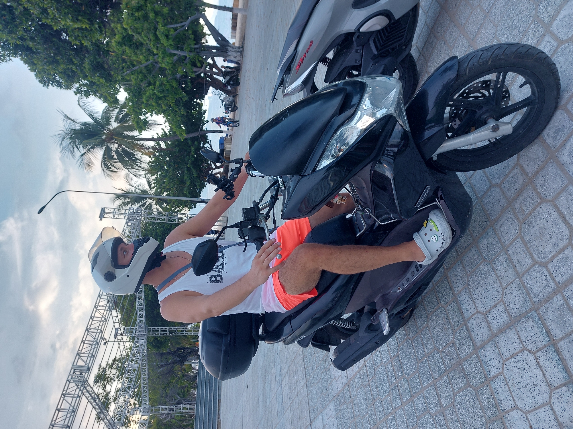 A person wearing a white tank top, orange shorts, a white helmet, and grey Crocs is sitting on a black scooter. The scooter has a black storage box on the back. The person is looking to their right. The scooter is parked on a paved area with a pattern of interlocking grey hexagonal tiles. In the background, there is a metal structure, palm trees, and other trees. The ocean is visible in the distance. Another scooter is parked to the right of the main scooter. The sky is cloudy. The scene appears to be in Nha Trang Ward, Vietnam, possibly along a beachfront promenade.