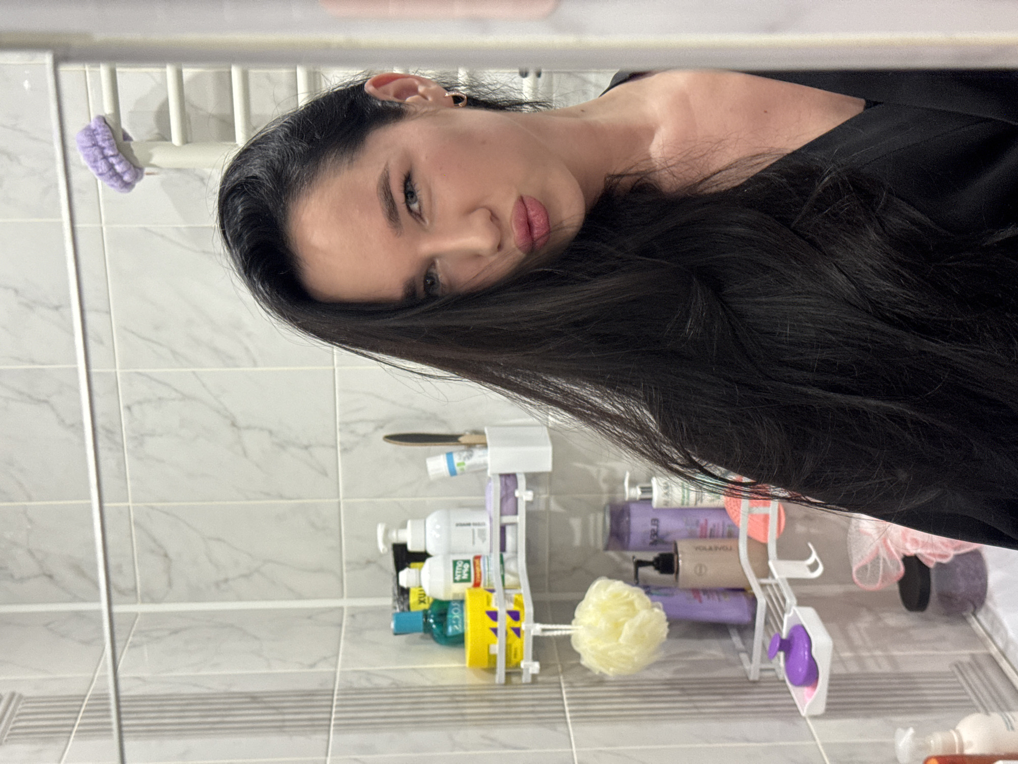 A young woman with long, dark hair and light skin poses for a selfie in a bathroom. She is wearing a black robe and has a playful expression, puckering her lips towards the camera. The bathroom features white tiled walls with a marble pattern. A white corner shelf unit is visible, stocked with various toiletries, including bottles of shampoo and lotion, a yellow container, and a purple brush. A yellow loofah hangs from the shelf. The woman is positioned to the right of the frame, with her face slightly turned towards the mirror, suggesting she is taking the photo using a reflection. The overall scene is well-lit, typical of an indoor bathroom environment. The location of Novosibirsk, Russia, is not explicitly identifiable from the visual information in the image alone.