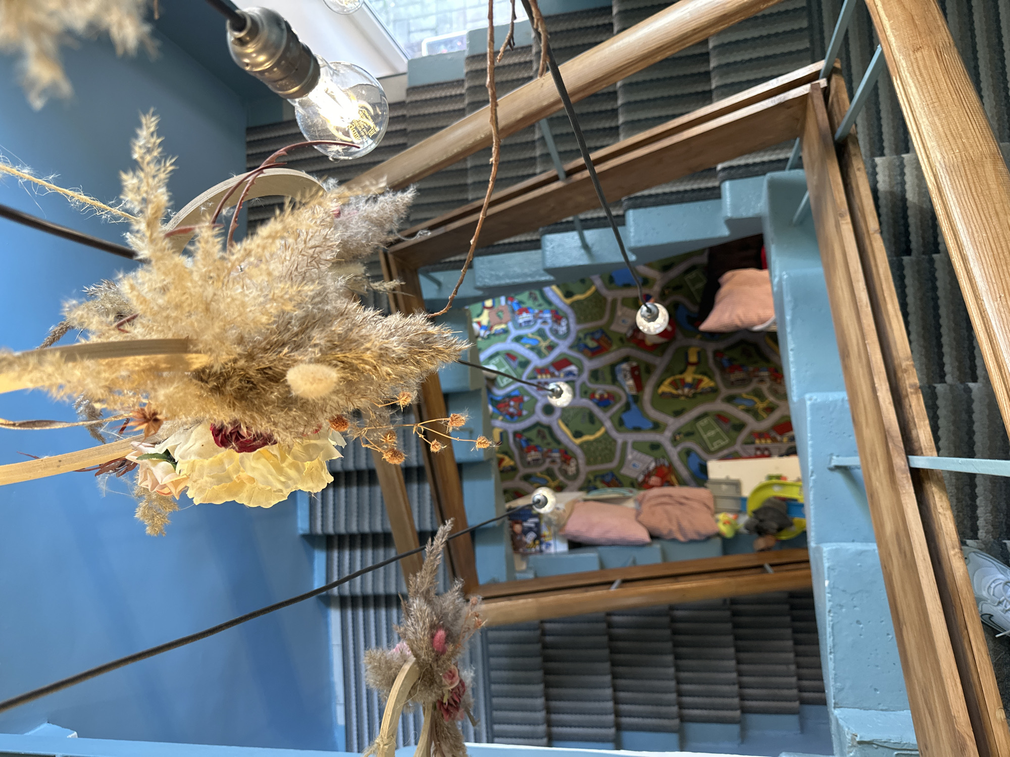 The image presents a low-angle perspective of a staircase within a building, likely in an urban setting, potentially Saint Petersburg, Russia, based on architectural styles and design elements common in that region. The foreground features decorative elements including a dried floral arrangement hanging from the ceiling with a visible lightbulb nearby. The staircase itself has steps painted in a light blue hue, contrasting with the striped pattern of the stair covering. The staircase is framed by wooden beams that form part of a decorative or structural element. In the background, through the openings created by the staircase and framing structure, a glimpse of a room is visible, showing a children's rug, pillows, and some toys. The image is well-lit, emphasizing the various textures and materials present in the scene.