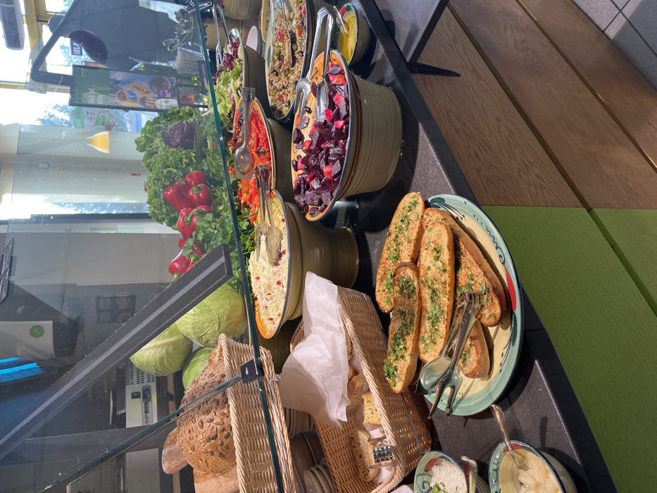 This image captures an indoor self-service food buffet in Općina Jasenice, Croatia, viewed from a patron's perspective. The foreground features a dark gray countertop displaying various food items. On the left, two small bowls contain a white food item (possibly a salad or rice) and diced yellow fruit, both with spoons. Centrally, a ceramic plate holds multiple slices of toasted garlic bread topped with green herbs, accompanied by serving tongs.
Further back, two wicker baskets are present: one filled with various bread rolls, including seeded varieties, and another containing slices of bread, some resting on white paper, both equipped with tongs. Behind a clear glass sneeze guard, a selection of prepared salads and vegetables is arranged in large ceramic bowls. These include a white salad with red seeds (possibly pomegranate), roasted red bell peppers, diced red beets, and a grain-based salad with olives and other vegetables, each with dedicated serving tongs. Fresh whole green cabbages, red bell peppers, assorted green leafy lettuces, and a dark red cabbage are displayed behind these prepared dishes. A smaller bowl of yellow dressing or oil is also visible.
The buffet counter's lower front paneling is green on the left and a wood-grain finish on the right. The background features large windows, reflecting outdoor light, a blurred human figure, a "MARCHÉ MENU" sign, and an internal green logo, suggesting a restaurant or cafeteria setting. No active individuals are directly interacting with the food.
