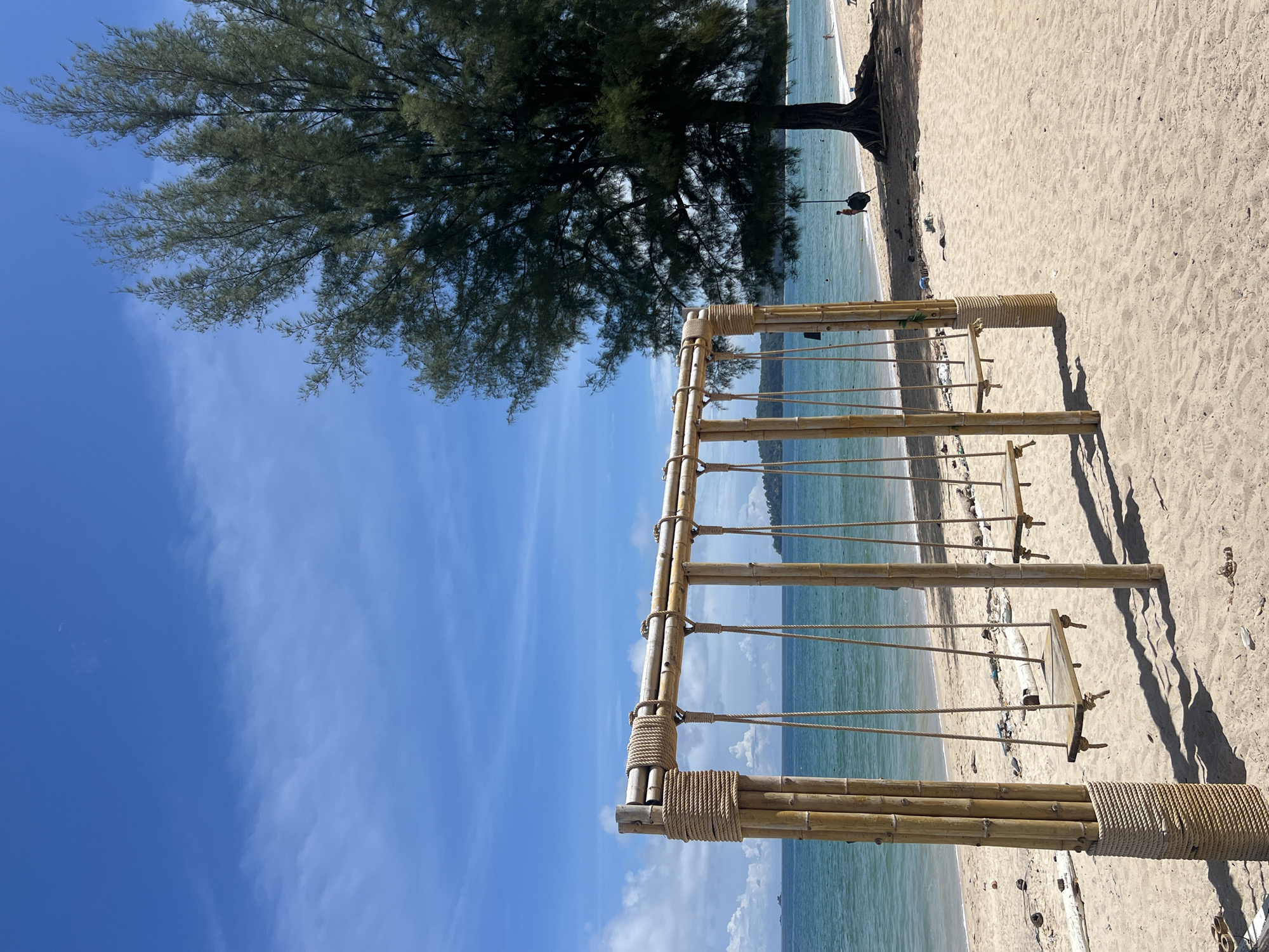 In Choeng Thale, Thailand, a wooden swing set constructed from bamboo poles is situated on a sandy beach. The swing set has four wooden seats suspended by ropes. To the right of the swing set, a large tree with dense foliage stands, its roots exposed. The ocean stretches out behind the beach, with calm turquoise water meeting a bright blue sky dotted with scattered white clouds. The sun casts shadows on the sand from the swings. In the distance, on the beach, a person is visible on a swing hanging from the tree, and other individuals are present in the water and on the shoreline. Debris, including driftwood and plastic, is scattered on the sand.