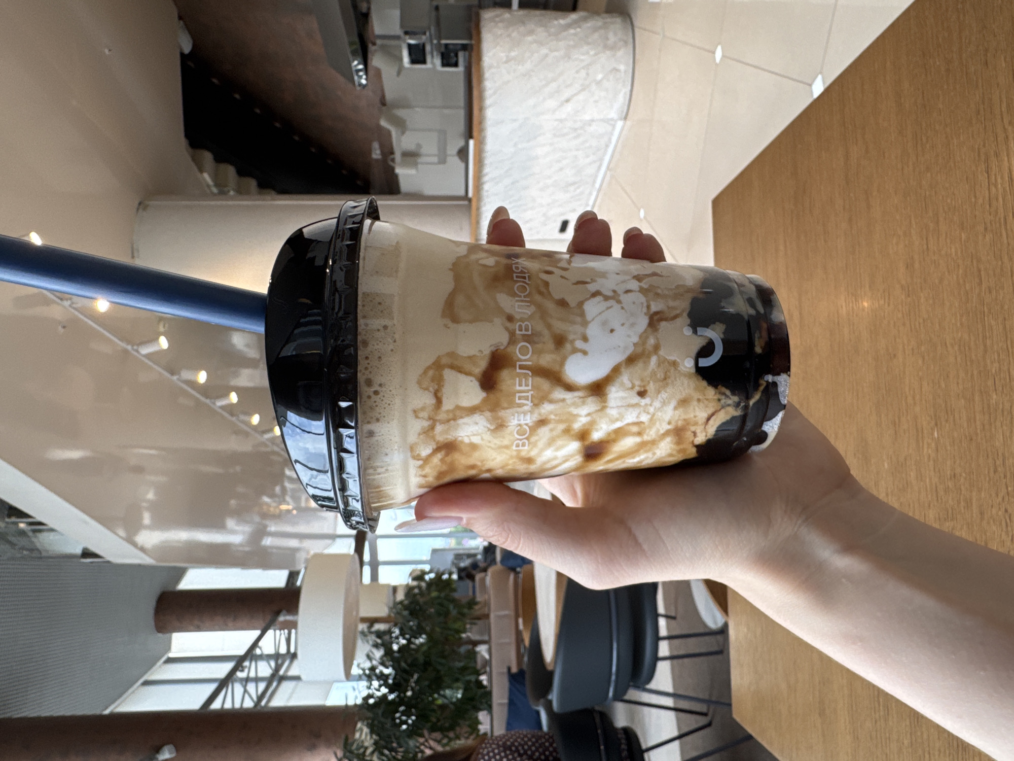 A hand holds a translucent plastic cup containing a brown-swirled beverage and black tapioca pearls, topped with a black lid and a blue straw. The text "ВСЕ ДЕЛО В ЛЮДЯХ" is visible on the cup. The background appears to be an indoor setting, possibly a cafe or restaurant, with large windows, seating areas, and a counter. The setting is consistent with the city of Yekaterinburg, Russia, given the Cyrillic text on the cup.