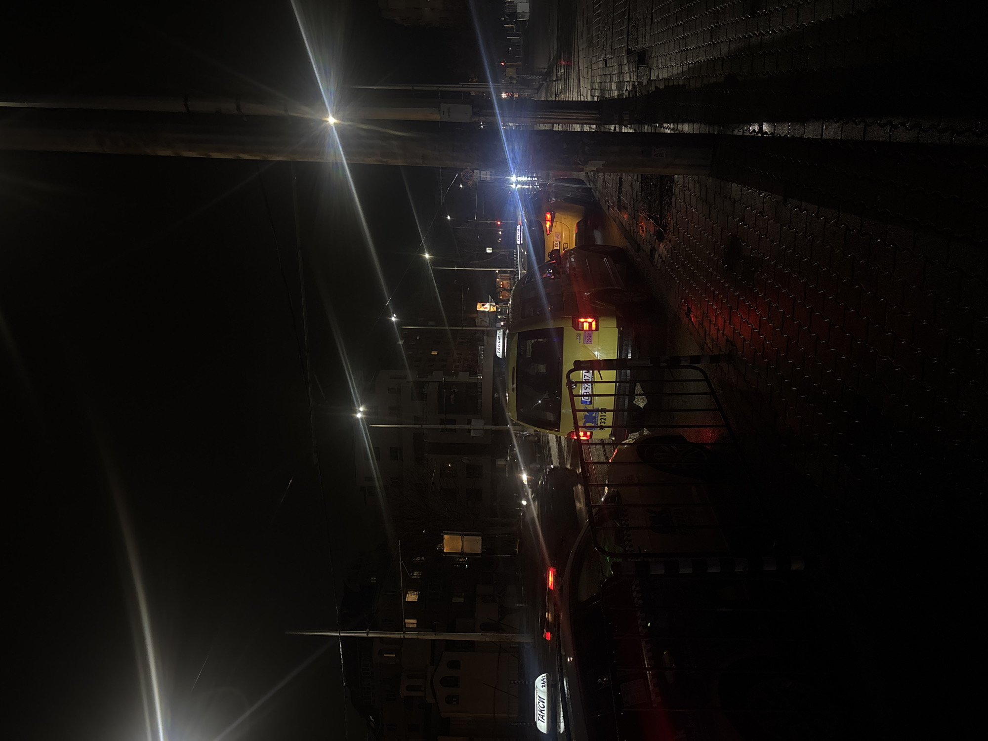 The image shows a street scene at night in Sofia, Bulgaria. A row of yellow taxis are parked along the right side of the street, facing forward, with their rear lights illuminated. Some of the license plates are visible, including one with Bulgarian text that reads "ТАКСИ" and "CB 9471." A sidewalk with a textured surface runs alongside the taxis. Streetlights are visible in the distance. The scene is dark, and the lighting is dominated by streetlights and vehicle headlights.
