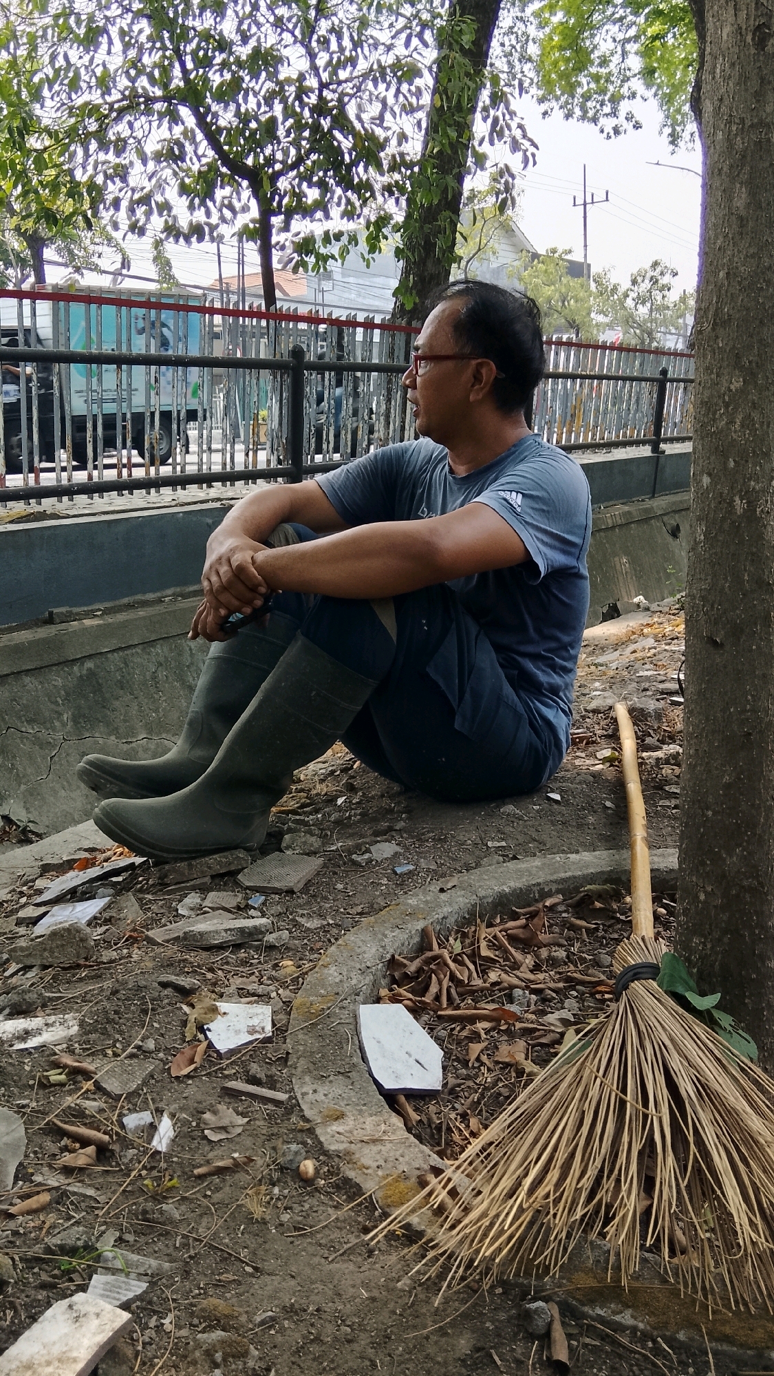 A man with glasses and dark hair is sitting cross-legged on the ground next to a tree in Gapurosukolilo, Indonesia. He is wearing a dark blue t-shirt, dark pants, and green rubber boots. His hands are clasped in front of him. A broom made of natural fibers lies on the ground to his right, leaning against a curved concrete structure. The ground is covered with dirt, dry leaves, and debris, including broken pieces of concrete and tile. In the background, a metal fence separates the area from a road where a truck and buildings can be seen. Trees with green leaves are present throughout the scene.