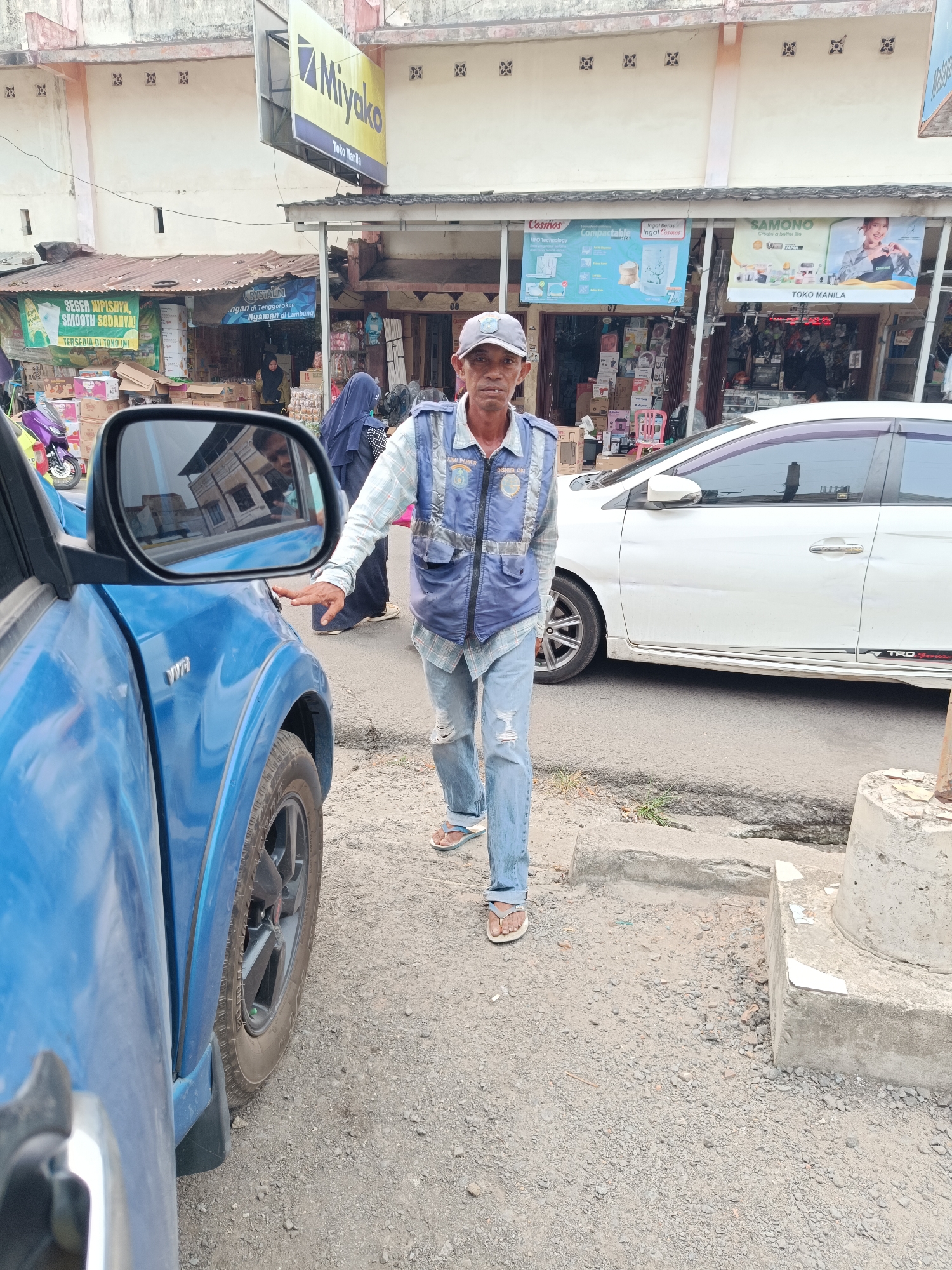 In Kayu Agung, Indonesia, a man stands by a blue vehicle. He wears a blue vest over a plaid shirt and ripped jeans, with sandals on his feet. He is looking towards the camera, with his right hand touching the side mirror of the blue car. Behind him, a white car is parked on a street lined with shops. The shops have signs advertising various products, including "Miyako" and "Cosmos." The scene appears to be a commercial area with pedestrian and vehicular traffic.